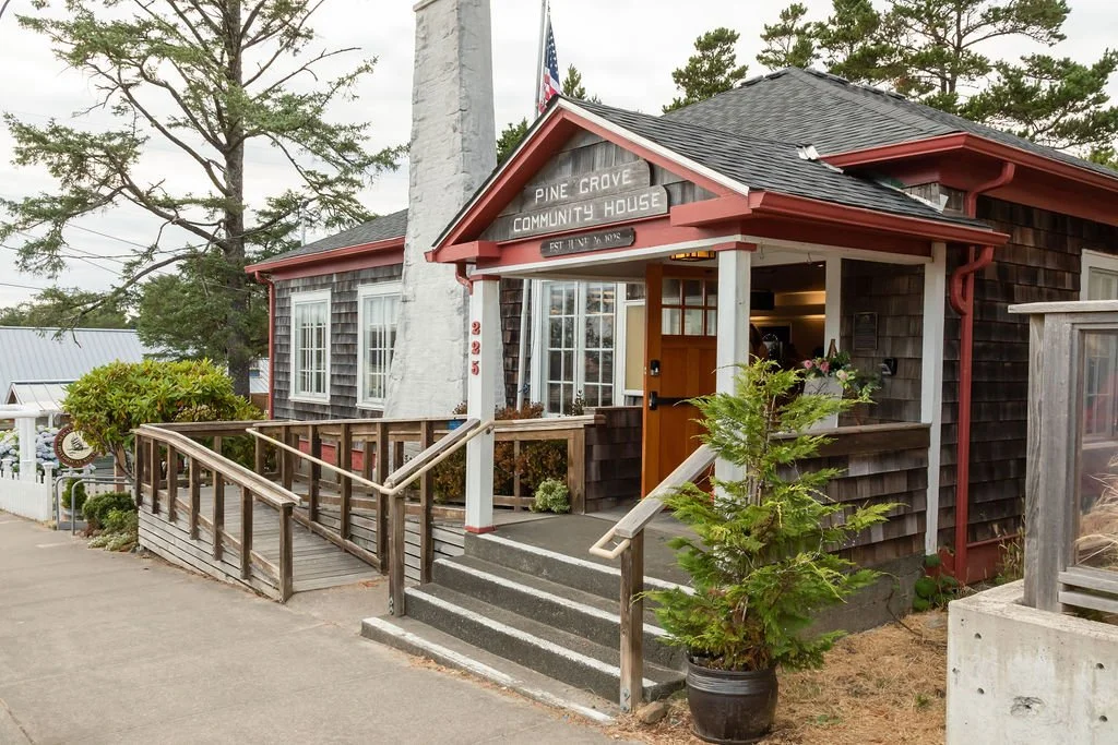 Small wooden community house with steps and ramp, labeled 'Pine Grove Community House,' with an American flag and greenery around.