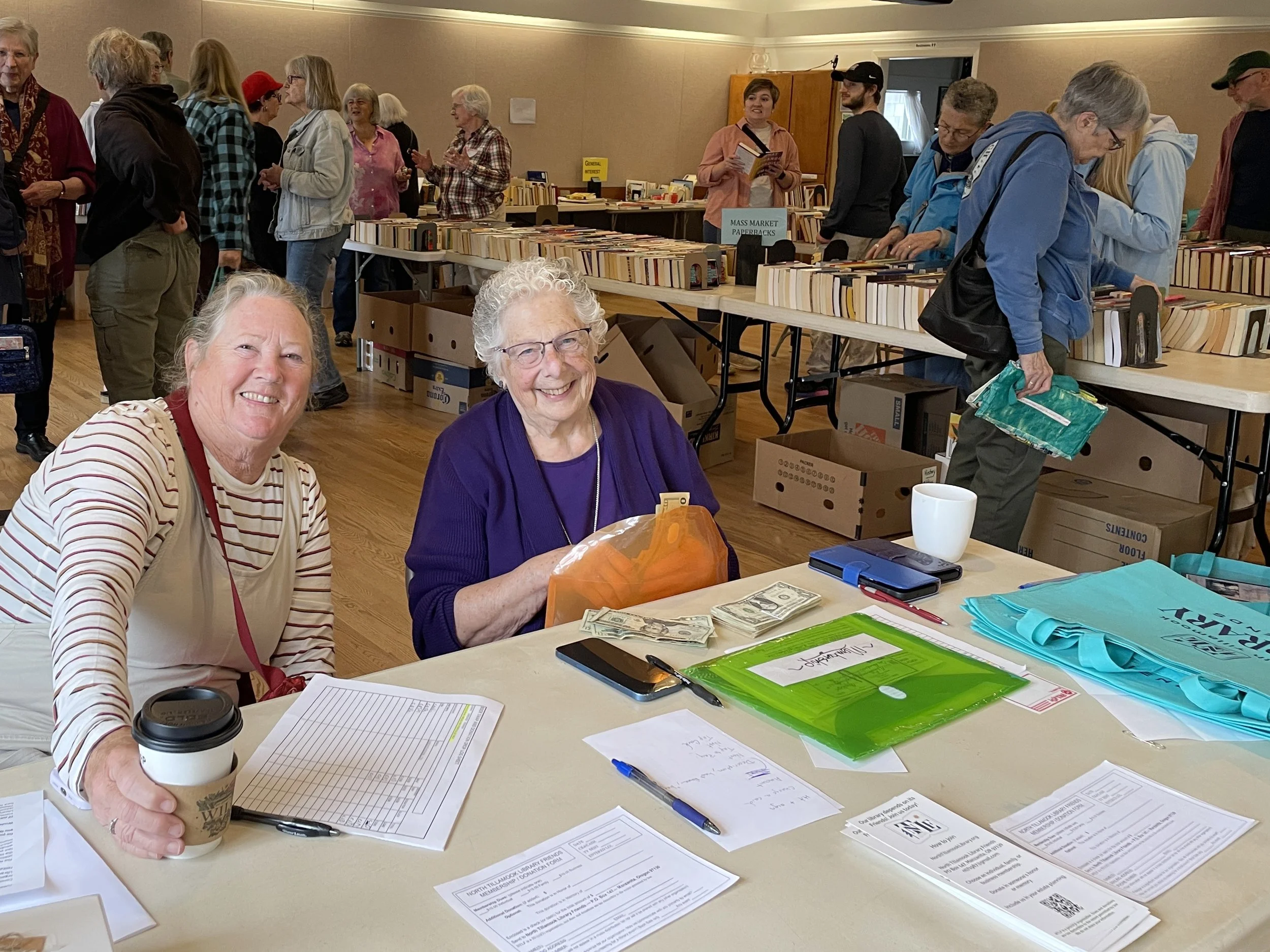 Two smiling women at a table with a stack of cash, papers, and a coffee cup during a book sale or market event, with people browsing in the background.