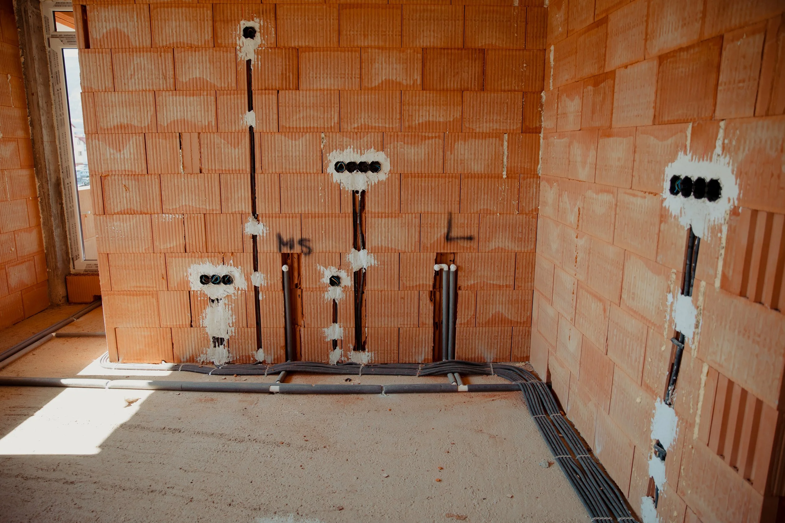 Innenansicht eines unfertigen Gebäudes mit unsymmetrischen bräunlichen Ziegelwänden, Steckdosen und Kabeln auf dem Boden und an der Wand, einige Kabel verlaufen an der Wand entlang, auf der linken Seite befindet sich ein Fenster.