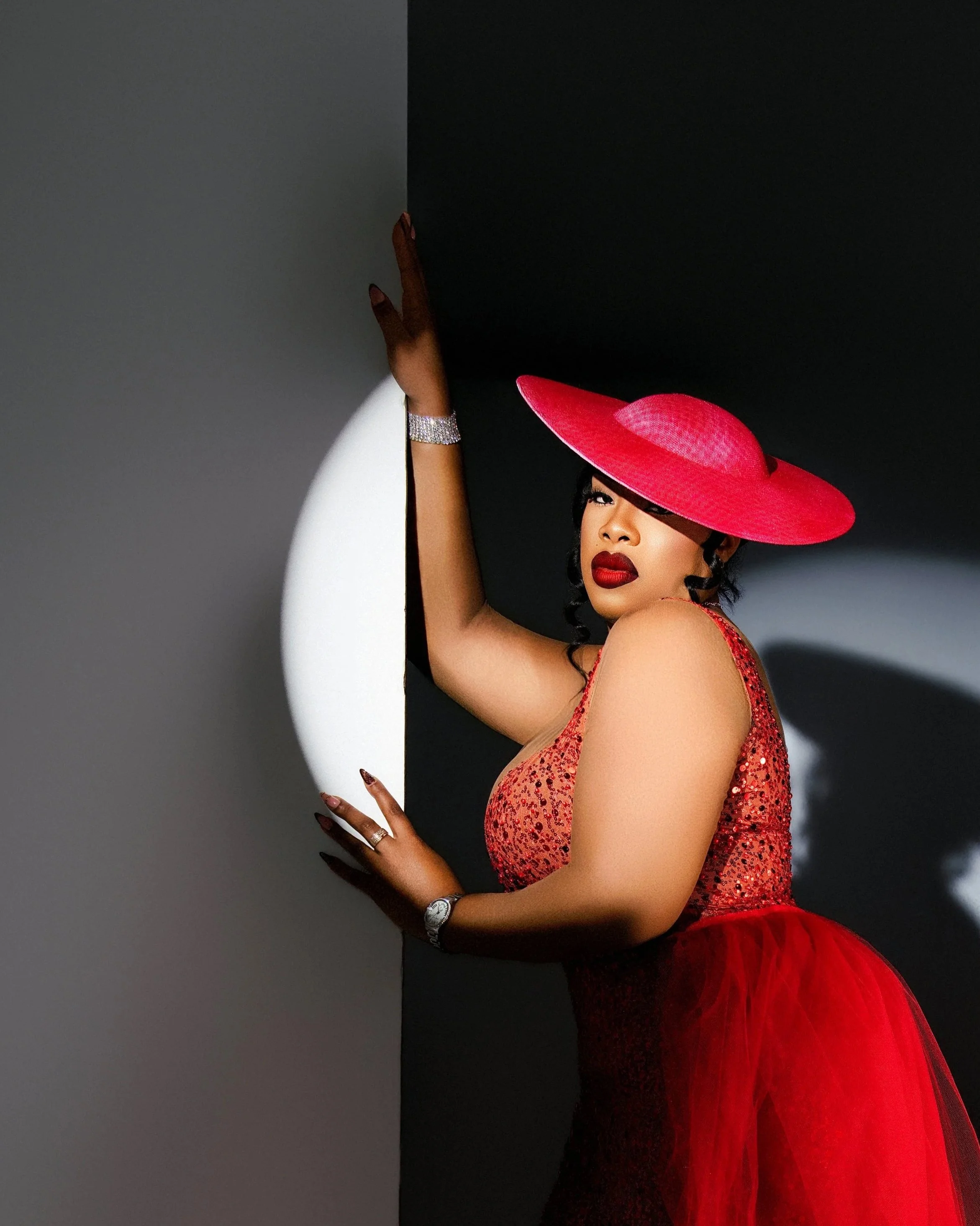 A bold editorial studio portrait photographed at Tonya Alexis Studios in Nassau, Bahamas, featuring a woman in a sparkling red sequin gown with a dramatic wide-brim fuchsia hat and rich red lipstick. Styled with luxury jewelry and posed against a mod