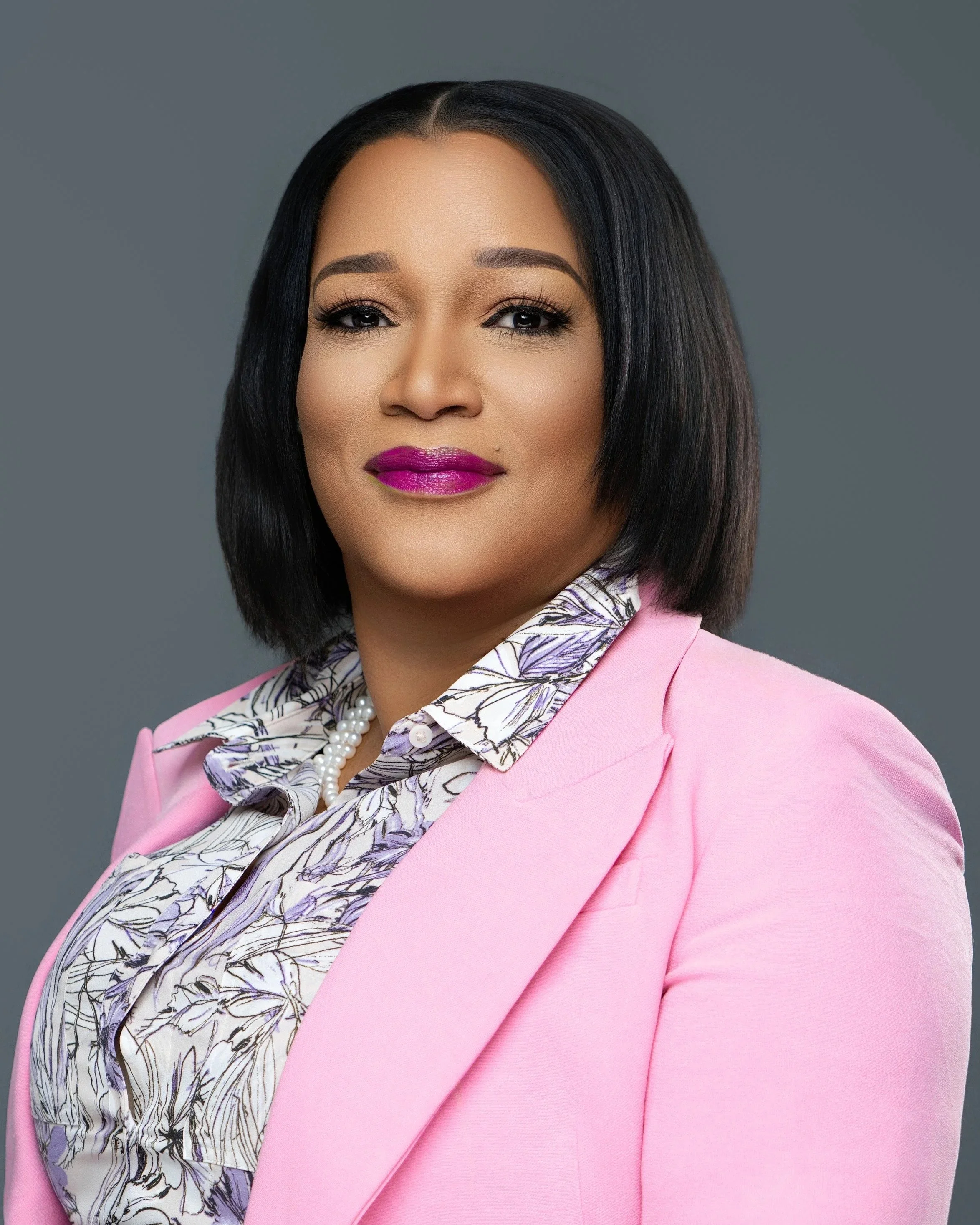 A vibrant professional headshot photographed at Tonya Alexis Studios in Nassau, Bahamas, featuring a confident woman styled in a soft pink blazer with a patterned blouse, pearl accessories, and polished makeup. Captured against a clean gray studio ba