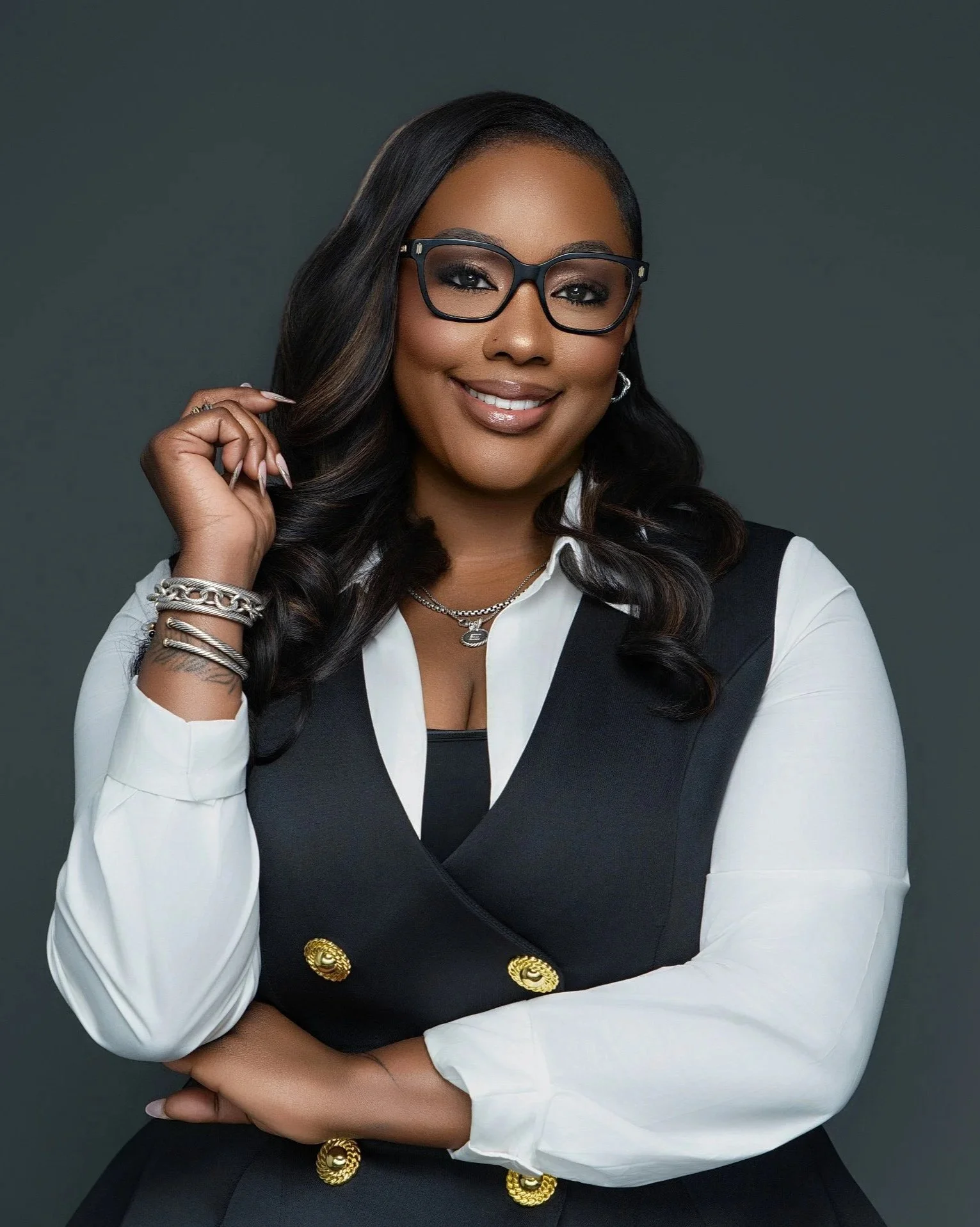 A polished personal branding portrait photographed at Tonya Alexis Studios in Nassau, Bahamas, featuring a confident woman wearing stylish black eyeglasses with a crisp white blouse and tailored black vest, posed with arms crossed. Set against a clea