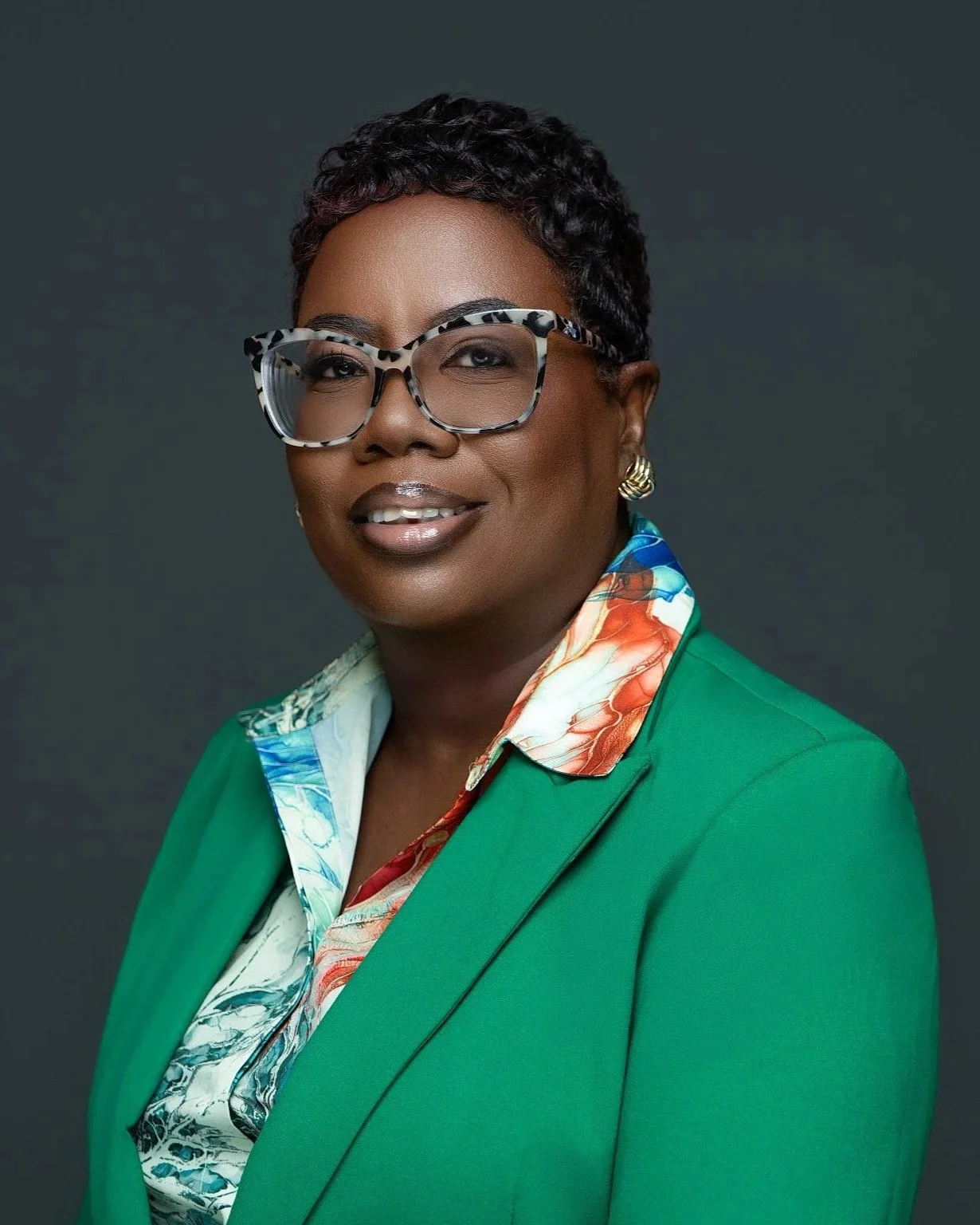 A polished professional headshot photographed at Tonya Alexis Studios in Nassau, Bahamas, featuring a confident woman in a vibrant green blazer with a colorful patterned blouse and stylish statement eyeglasses. Set against a clean dark studio backgro