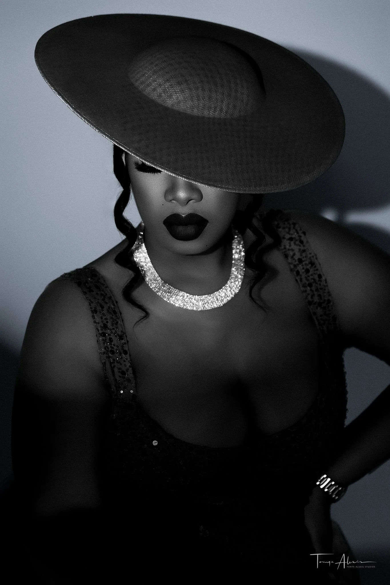 Black-and-white editorial studio portrait of a woman in a wide-brim hat and sequin dress with a sparkling necklace, photographed at Tonya Alexis Studios in Nassau, Bahamas.