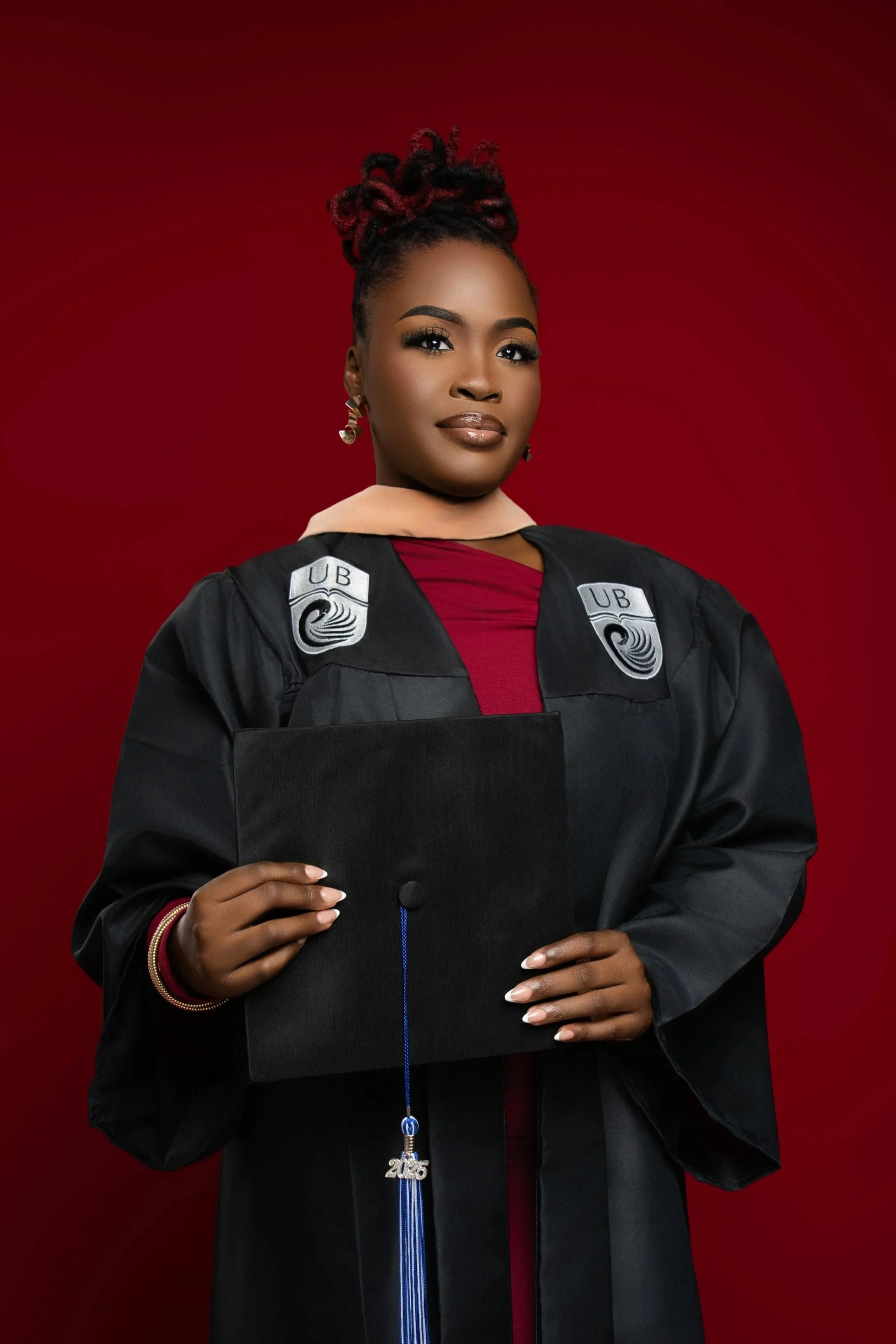 Professional university graduation portrait in Nassau, Bahamas by Tonya Alexis Studios, featuring a confident female graduate in a black academic gown holding a graduation cap against a deep red studio backdrop.