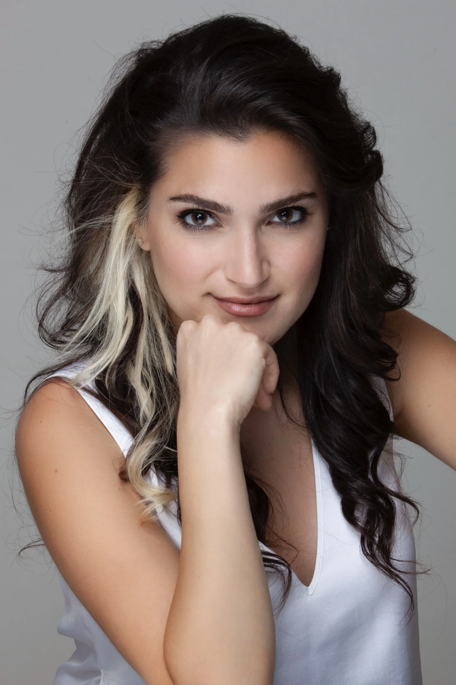A young woman with dark and blonde wavy hair, wearing a white sleeveless top, resting her chin on her hand and looking into the camera against a plain gray background.