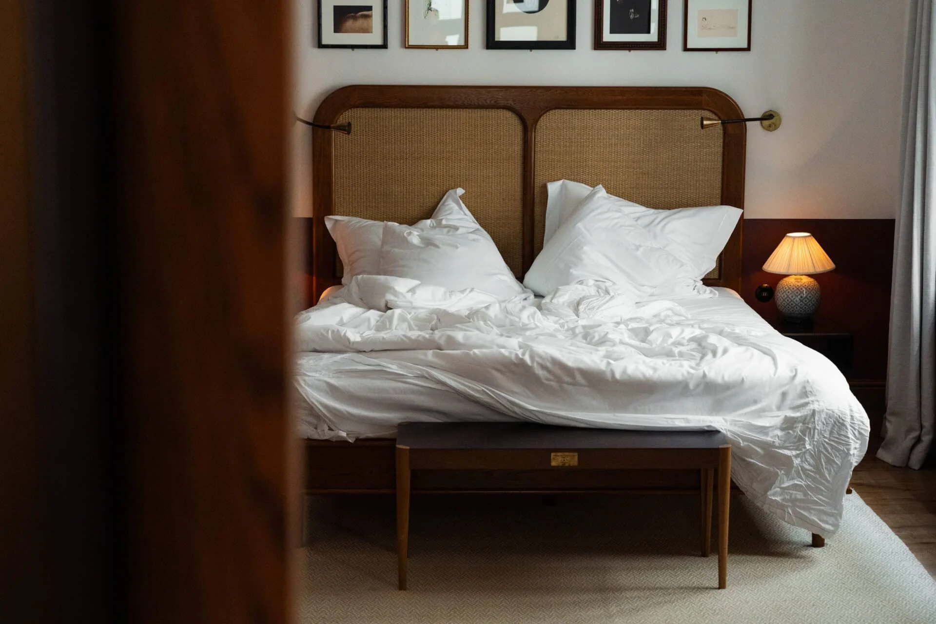 Cozy boutique luxury hotel bedroom featuring an unmade bed with white linens, a woven rattan headboard, warm bedside lighting, and elegant vintage-inspired decor.