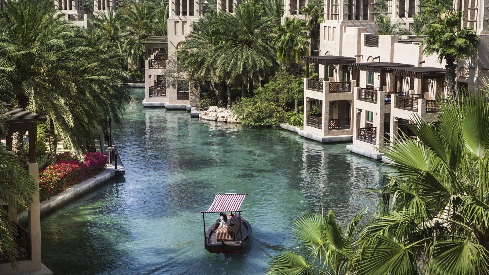 Jumeirah Dar Al Masyaf in Dubai featuring serene turquoise canals, palm-lined pathways, and traditional Arabian summerhouse villas with an abra boat gliding through the water—luxury waterfront resort experience.