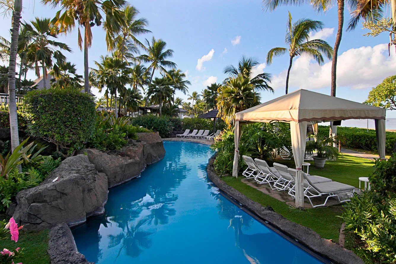 Beautiful tropical resort pool in Maui with a winding lazy river, lush palm trees, and shaded cabanas surrounded by vibrant greenery.