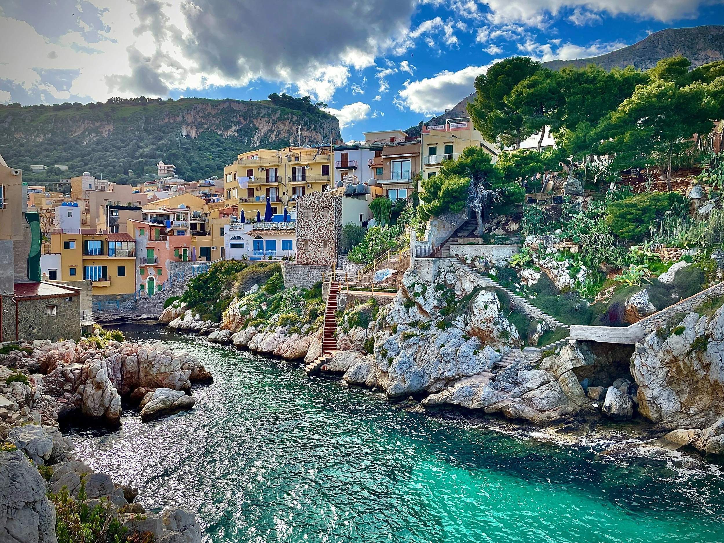 Colorful coastal village with stone stairways and turquoise water along the rocky shoreline in Sicily, Italy.