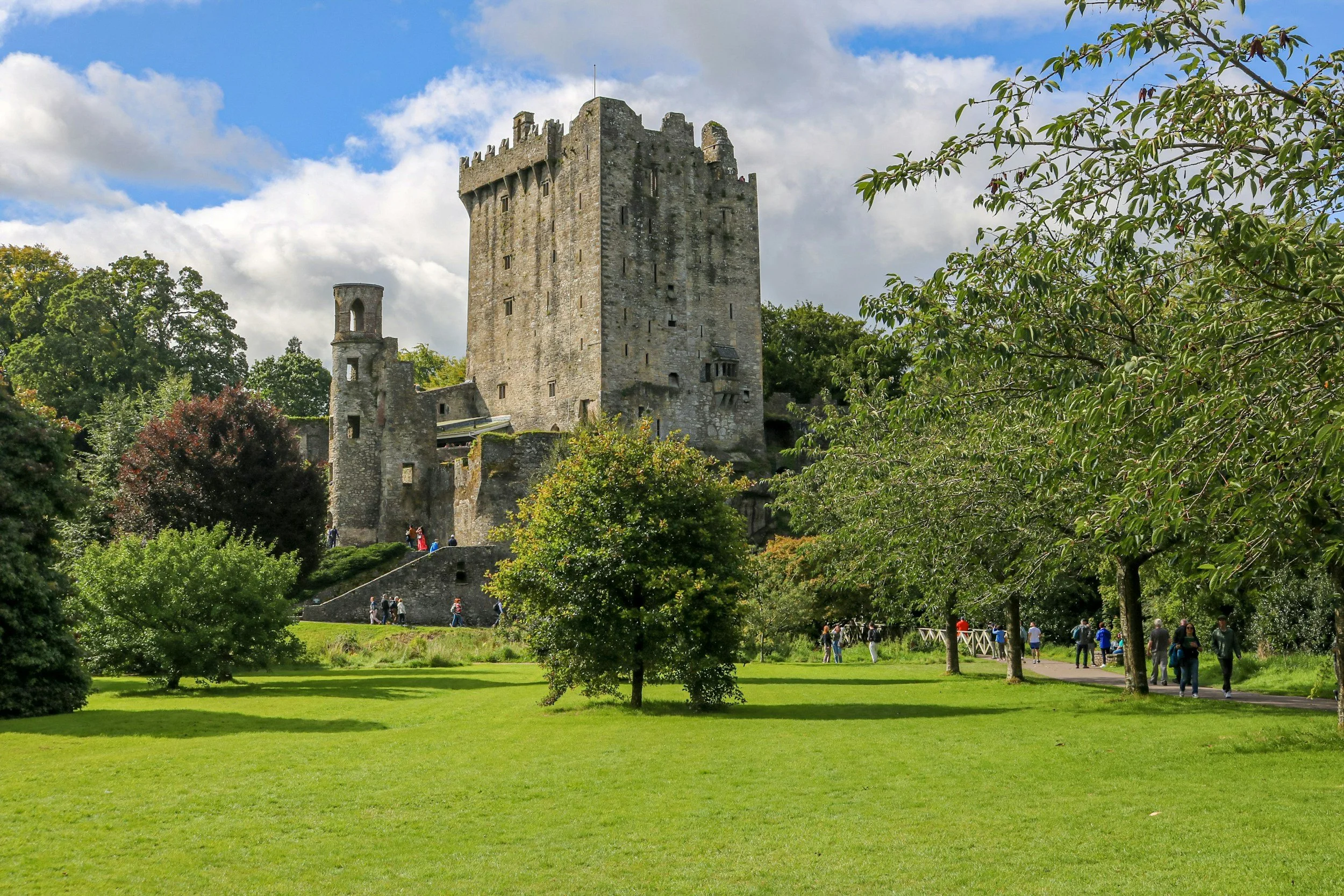 Blarney Castle in County Cork, Ireland surrounded by lush green gardens and walking paths under a bright blue sky.
