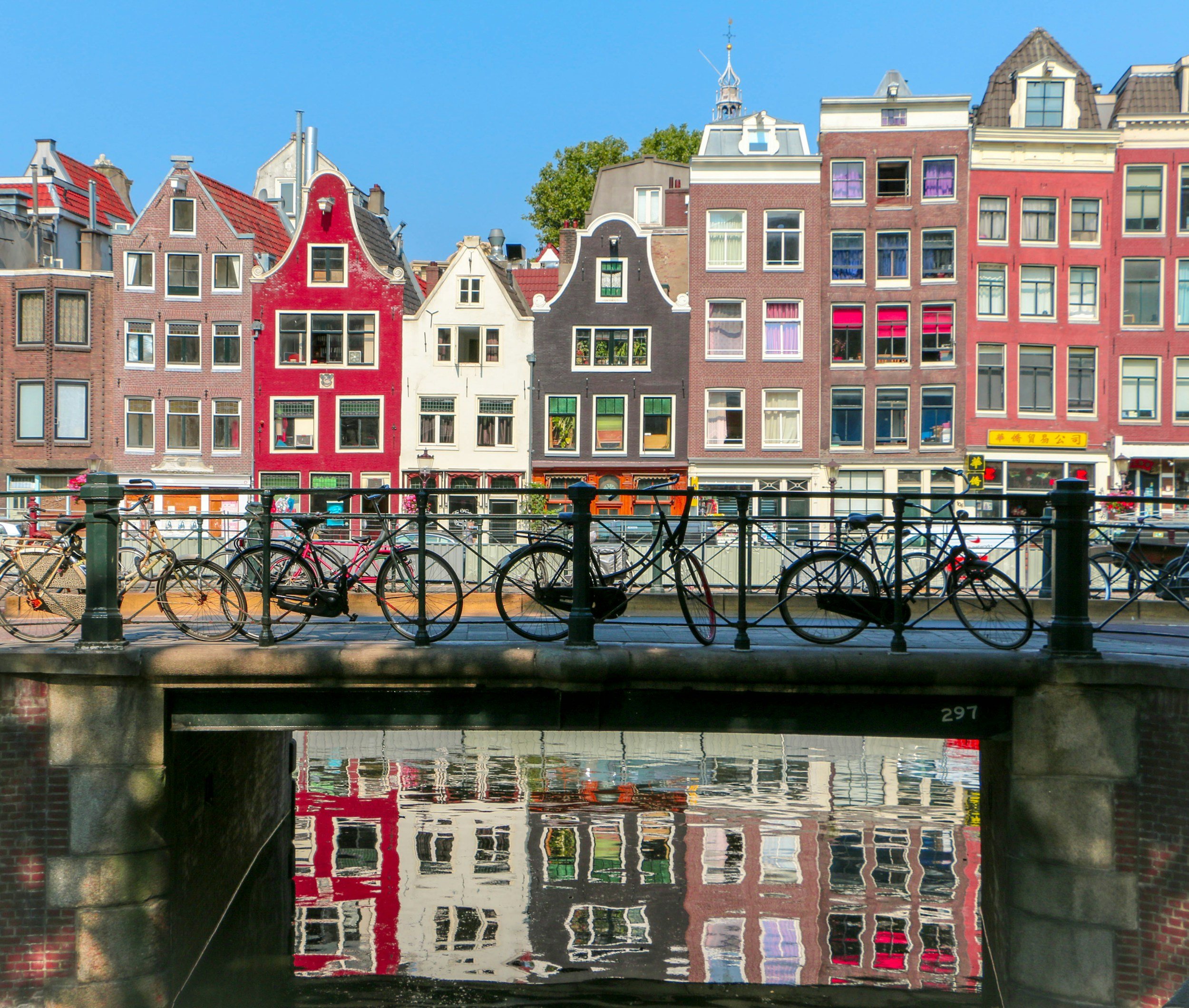 Colorful canal houses in Amsterdam with bicycles lined along a bridge and reflections in the water below.