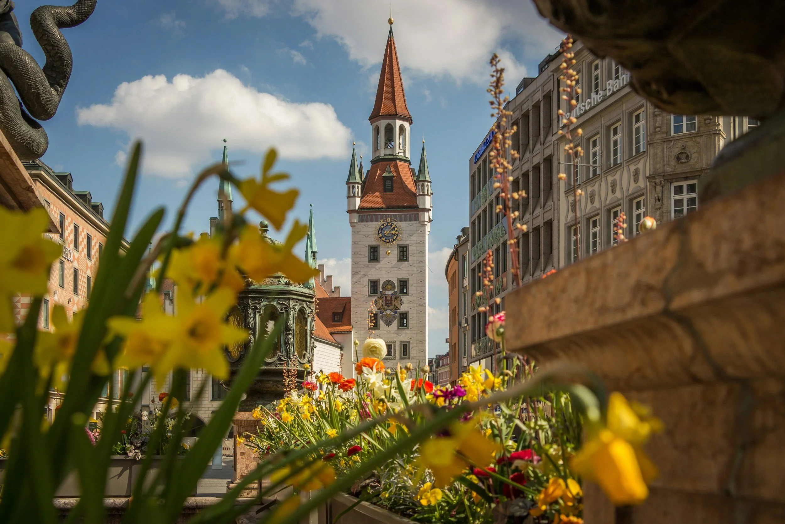 Historic Old Town Munich with colorful spring flowers in the foreground and the iconic Old Town Hall clock tower under a blue sky.