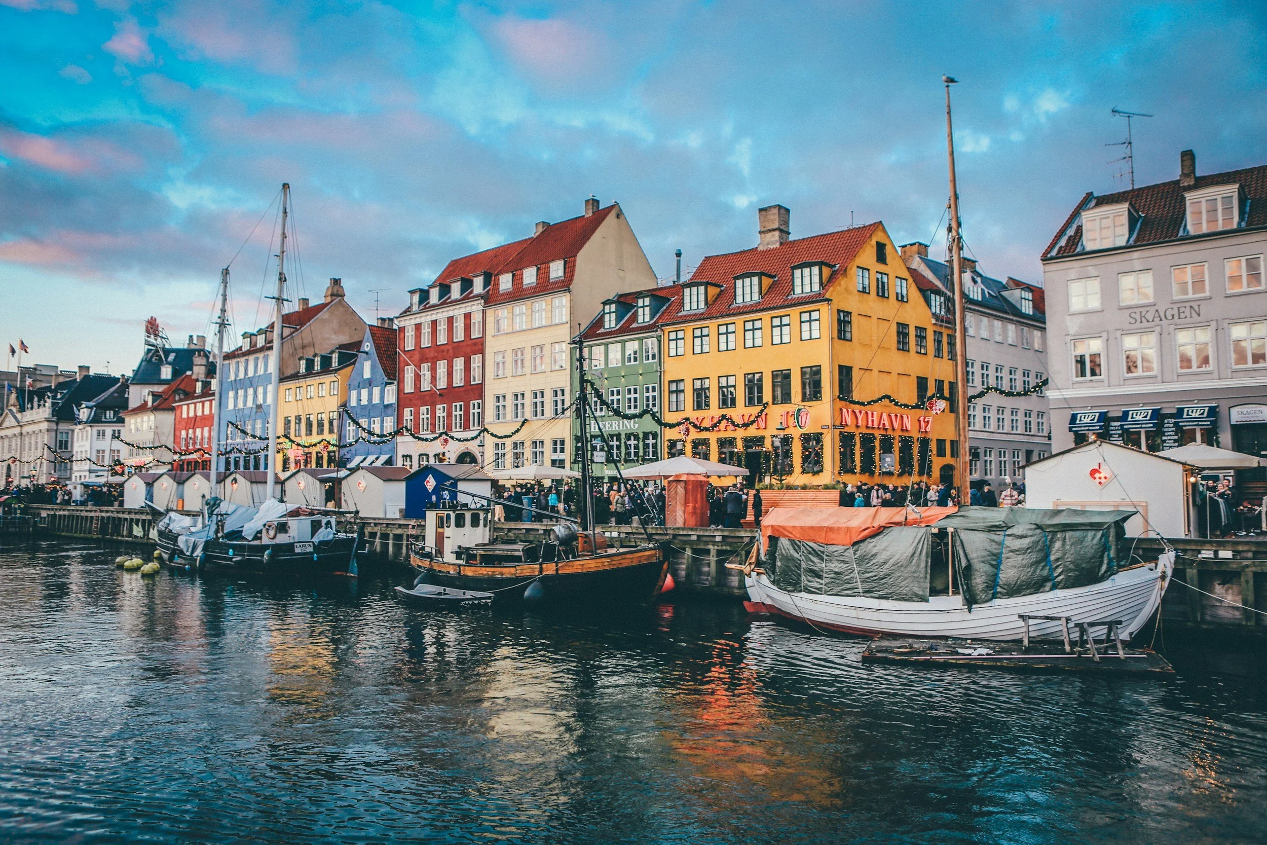 Colorful waterfront buildings along Nyhavn canal in Copenhagen with boats, reflections in the water, and lively atmosphere at sunset.