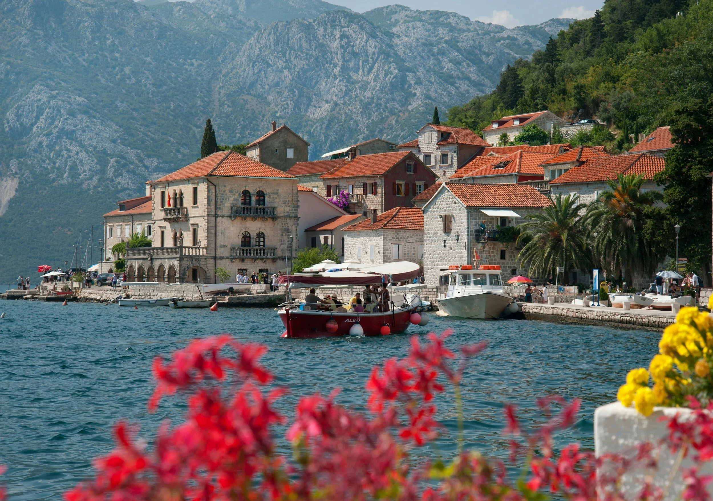 Waterfront view of Perast, Montenegro, featuring historic stone buildings with terracotta rooftops, colorful boats floating on the Bay of Kotor, and dramatic mountains rising in the background.