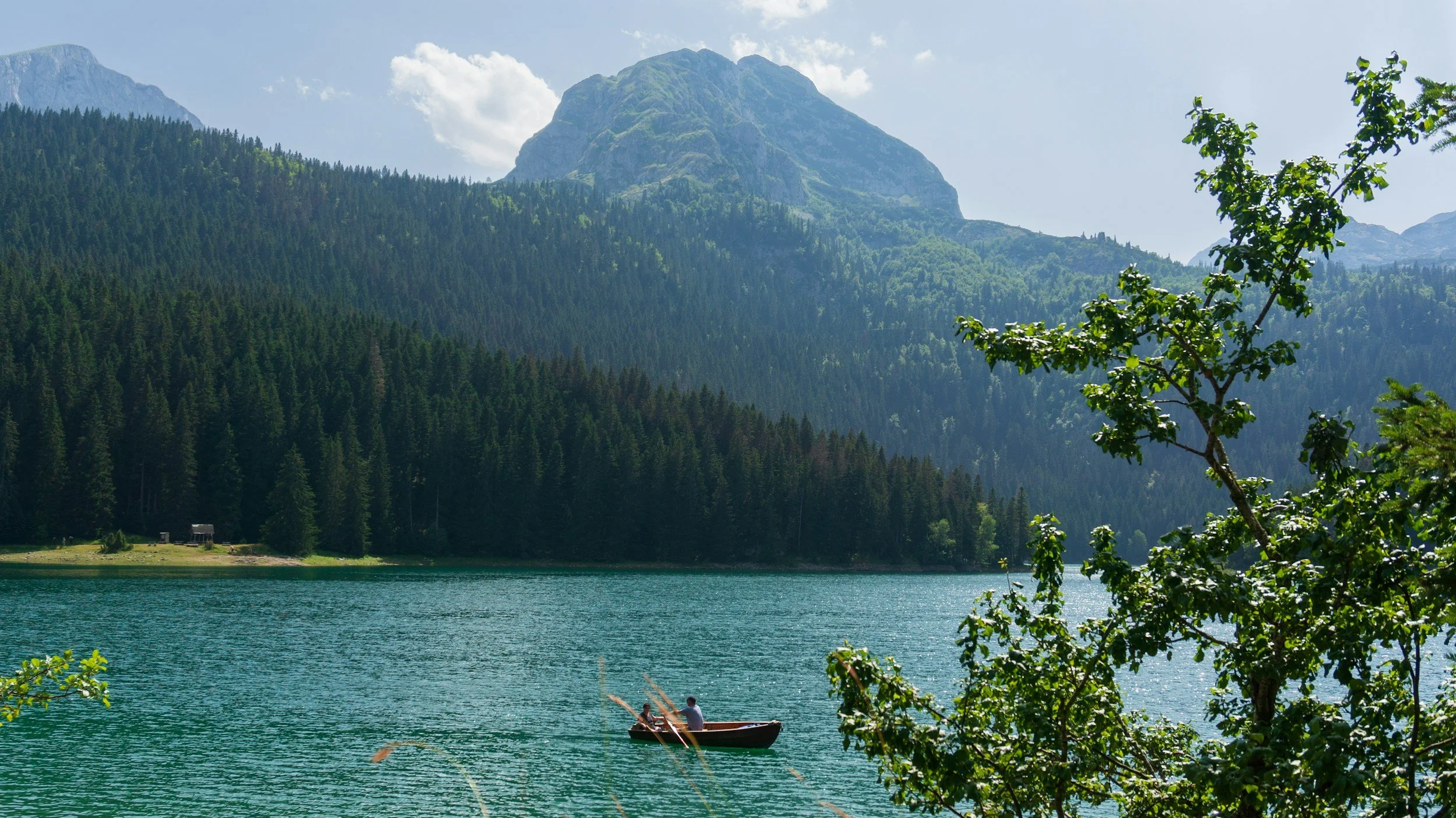 Small wooden boat floating on the turquoise waters of Black Lake in Durmitor National Park, Montenegro, surrounded by dense evergreen forests and dramatic mountain peaks on a sunny day.