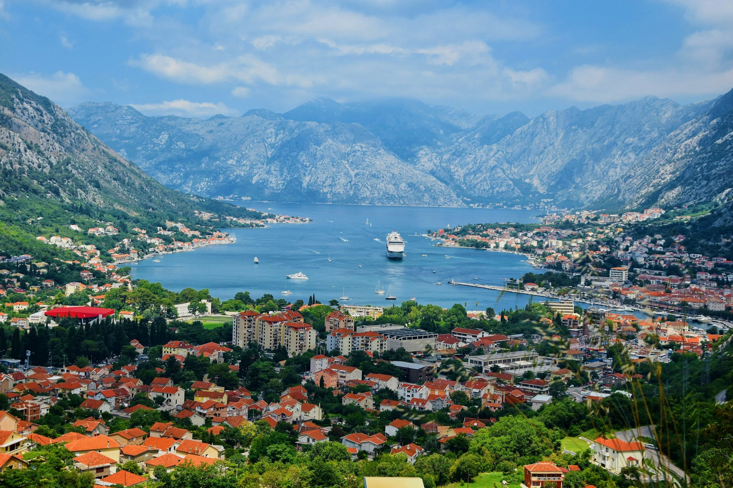 Panoramic view of the Bay of Kotor in Montenegro, featuring a cruise ship sailing through the fjord-like bay, charming red-roofed coastal towns, and dramatic limestone mountains beneath a cloudy sky.