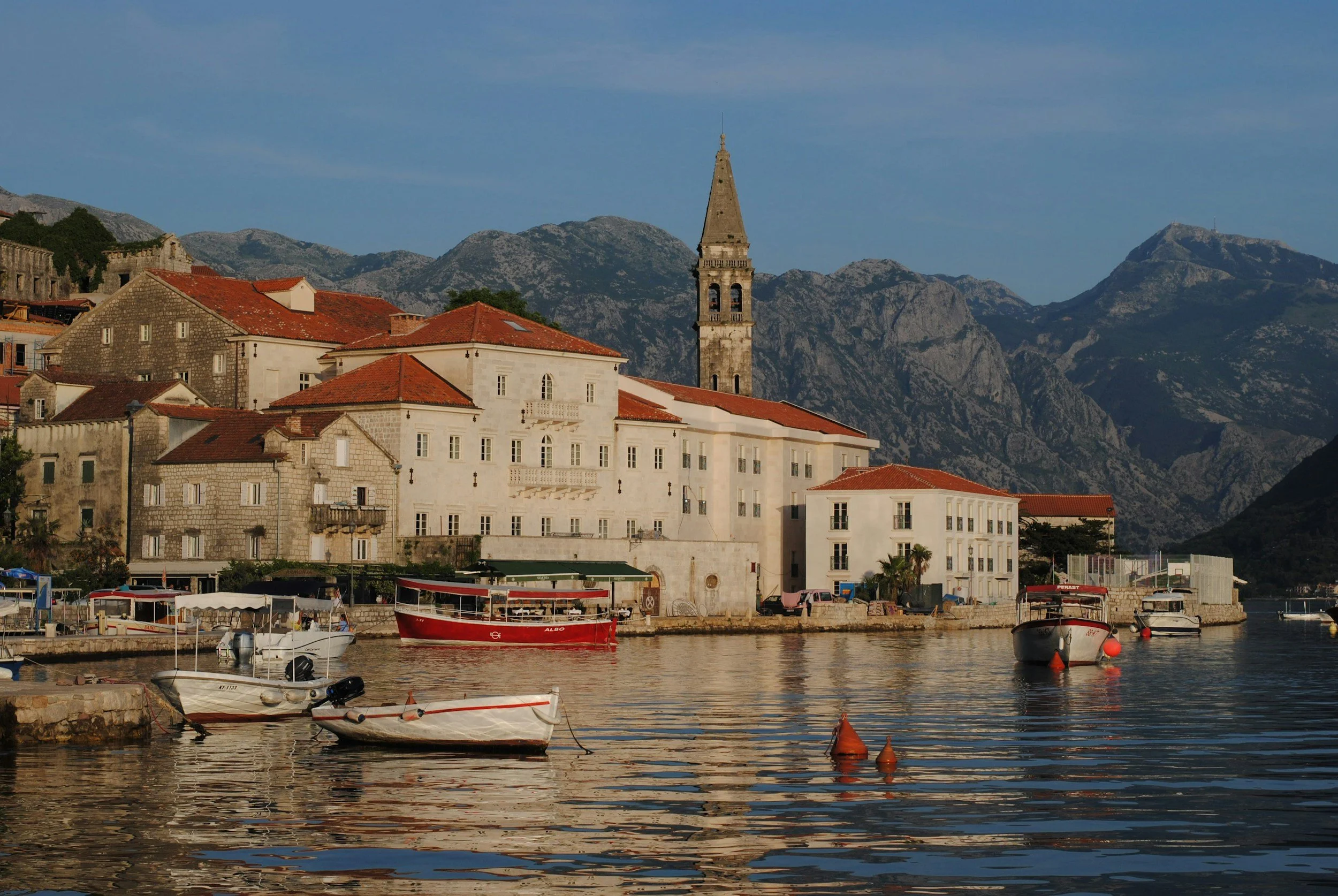 Historic waterfront village of Perast in Montenegro, featuring traditional stone buildings, a church bell tower, small boats floating in the Bay of Kotor, and dramatic mountain scenery in the background.