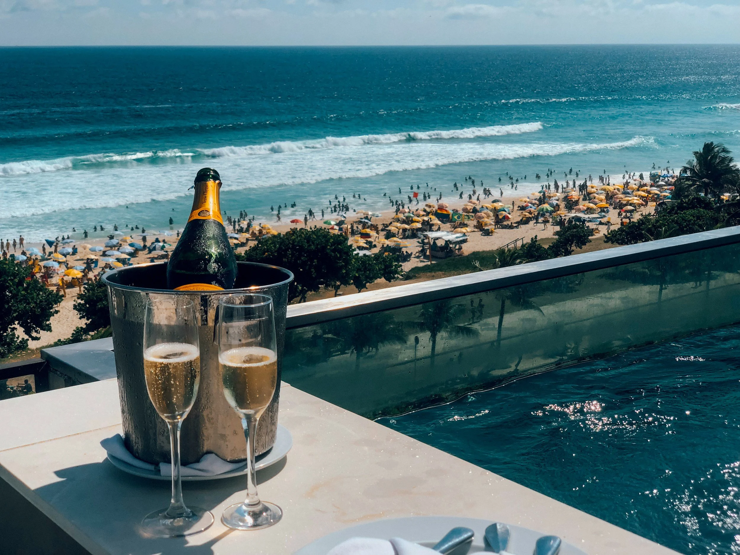 Champagne and two glasses overlooking a lively tropical beach and turquoise ocean from a luxury oceanfront pool terrace.
