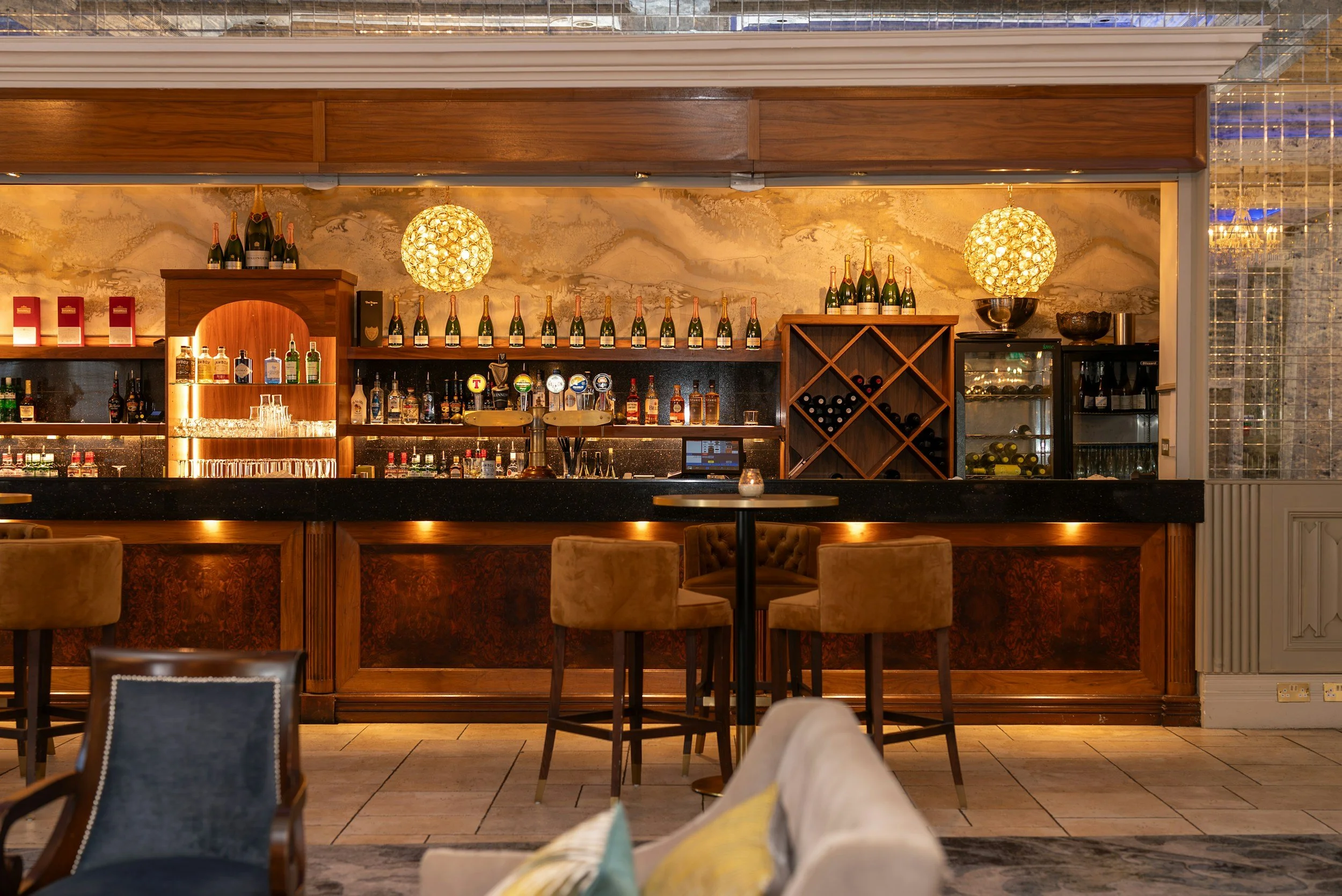 Elegant luxury hotel bar with warm wood finishes, glowing pendant lights, plush seating, and an extensive display of champagne, wine, and spirits.