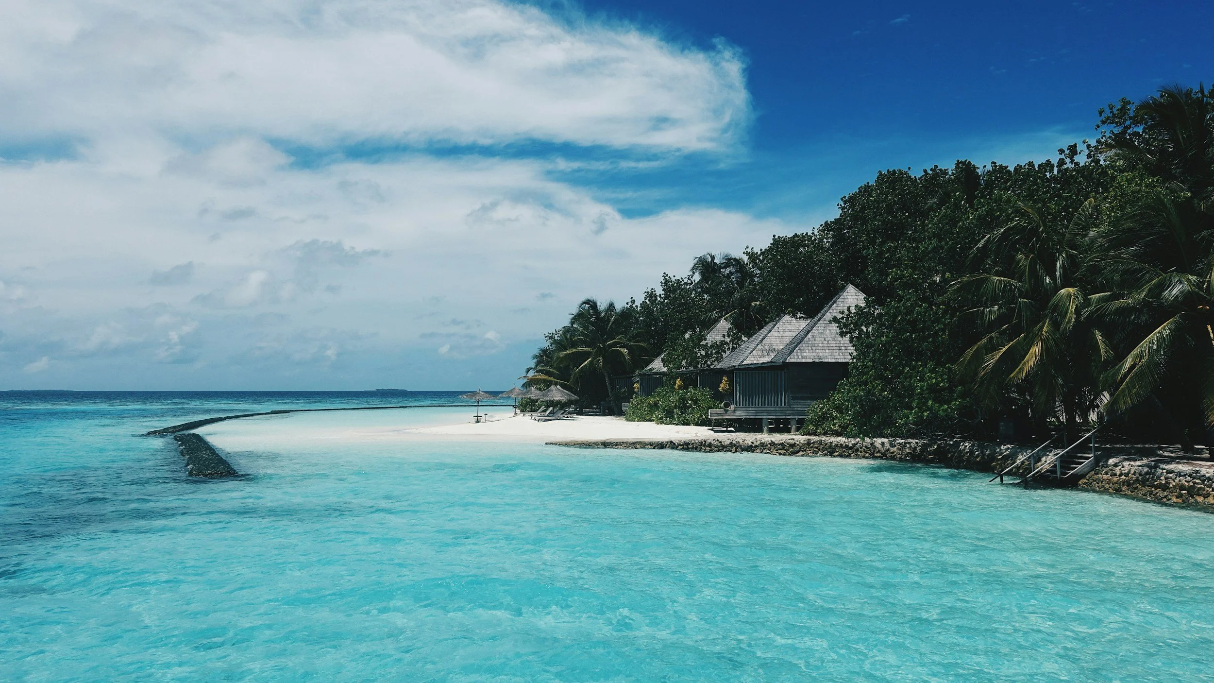 Luxury Maldives tropical island resort surrounded by palm trees, white sand, and crystal-clear turquoise water beneath a bright blue sky.