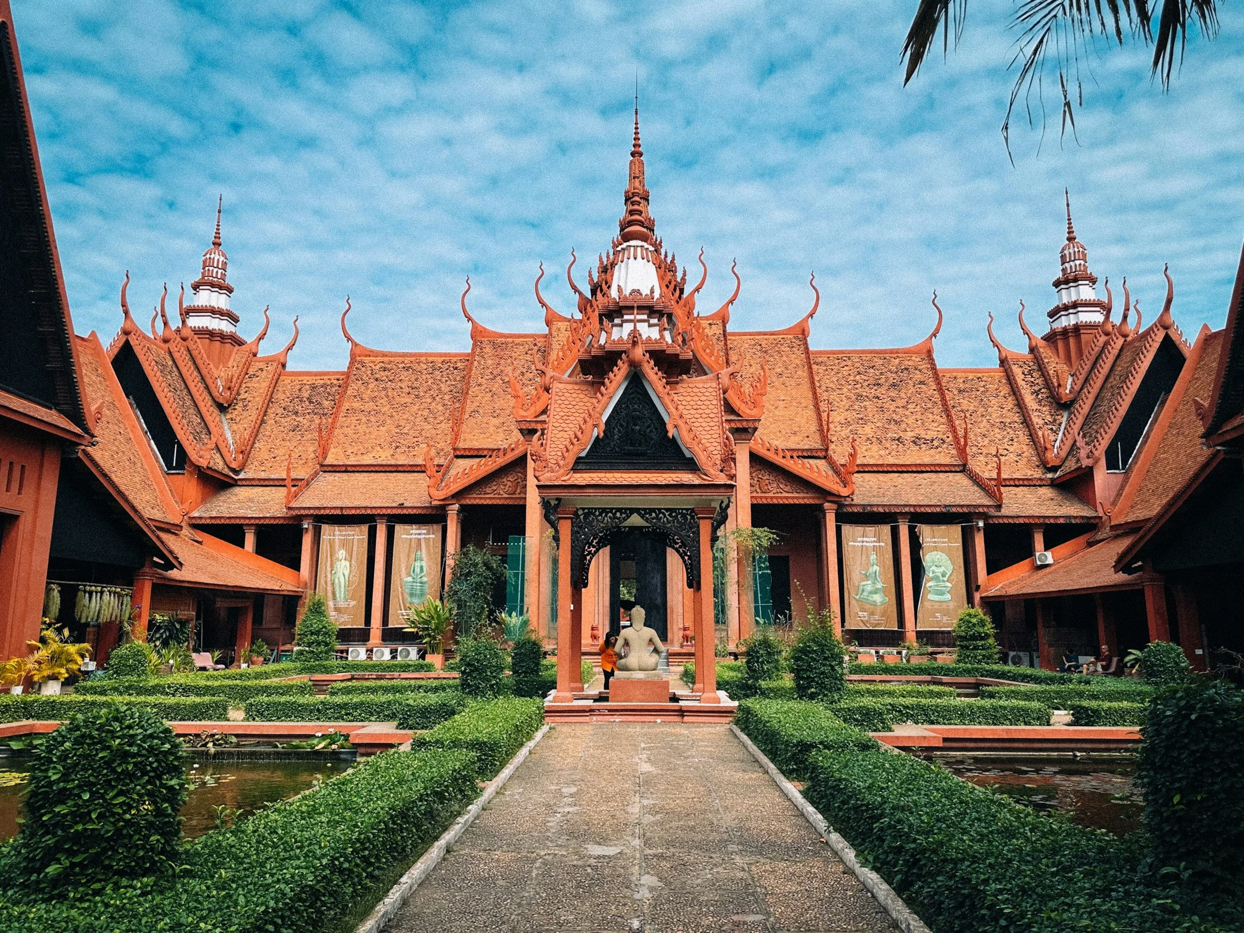 Traditional Khmer architecture at the National Museum of Cambodia in Phnom Penh, featuring ornate terracotta roofs, a central courtyard, lush gardens, and a stone sculpture.