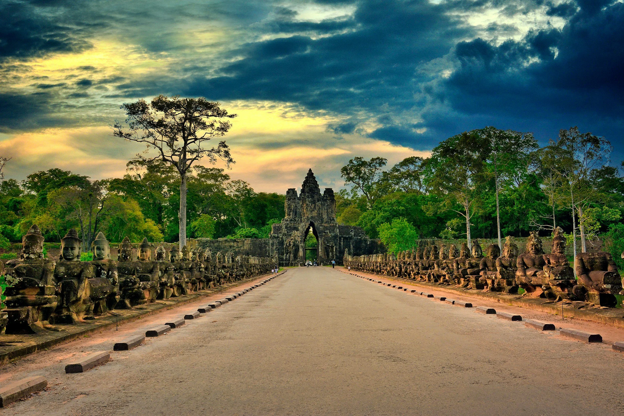 A dramatic view of the South Gate of Angkor Thom in Siem Reap, Cambodia, with rows of ancient stone statues lining the causeway beneath a colorful, cloud-filled sky.