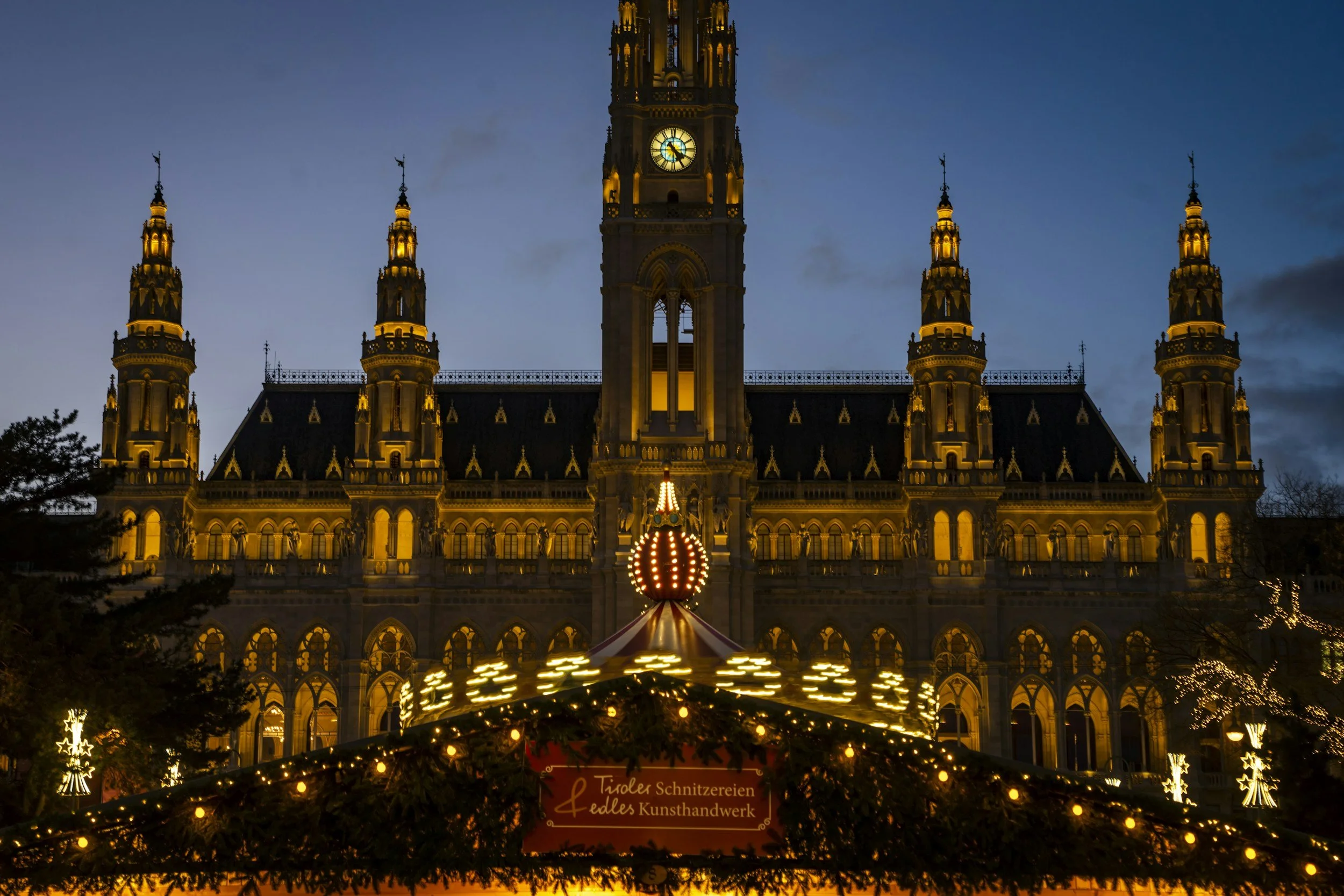 Vienna Christmas market at night featuring illuminated wooden holiday stalls, festive decorations, and the stunning Gothic architecture of Vienna City Hall (Rathaus) glowing against the evening sky.