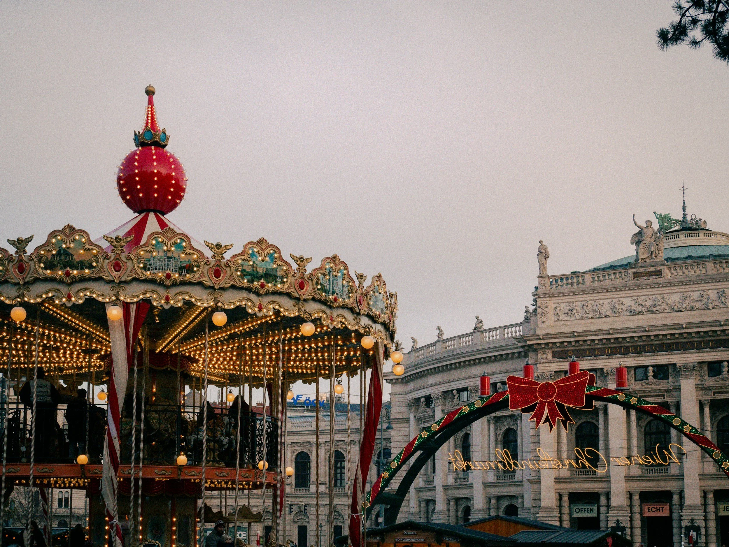Festive Christmas market in Vienna, Austria, featuring an illuminated vintage carousel, holiday decorations, and the historic Burgtheater building in the background.