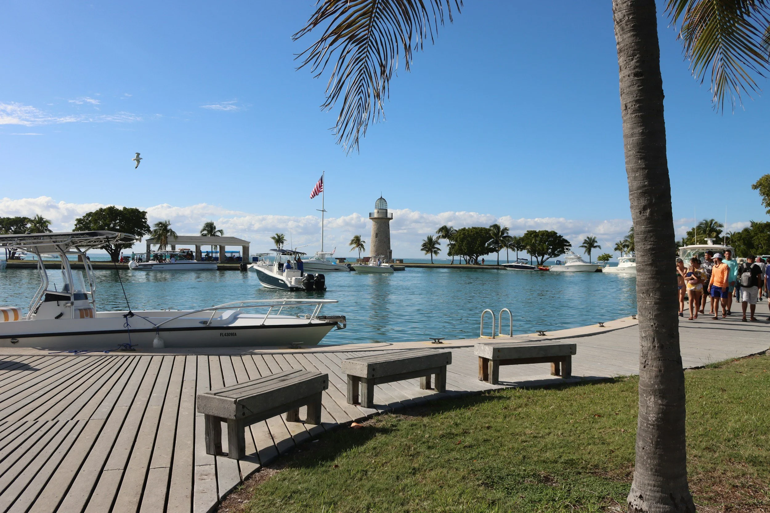 Scenic waterfront marina in Miami, Florida, featuring docked boats, a stone lighthouse, palm trees, and visitors strolling along the boardwalk on a sunny day.