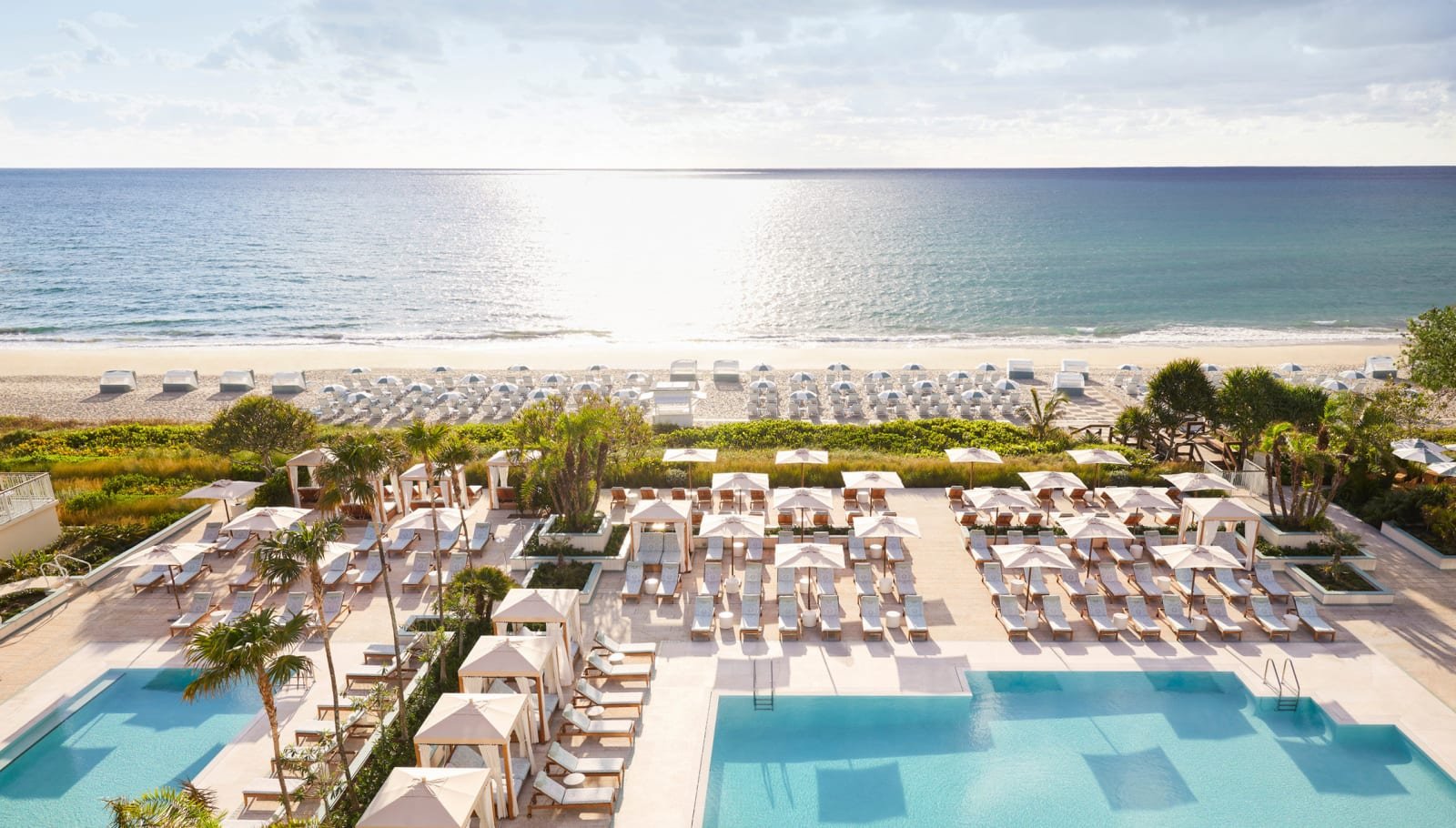 Aerial view of a luxury beachfront resort featuring turquoise swimming pools, private cabanas, white lounge chairs, and an expansive sandy beach overlooking the ocean.