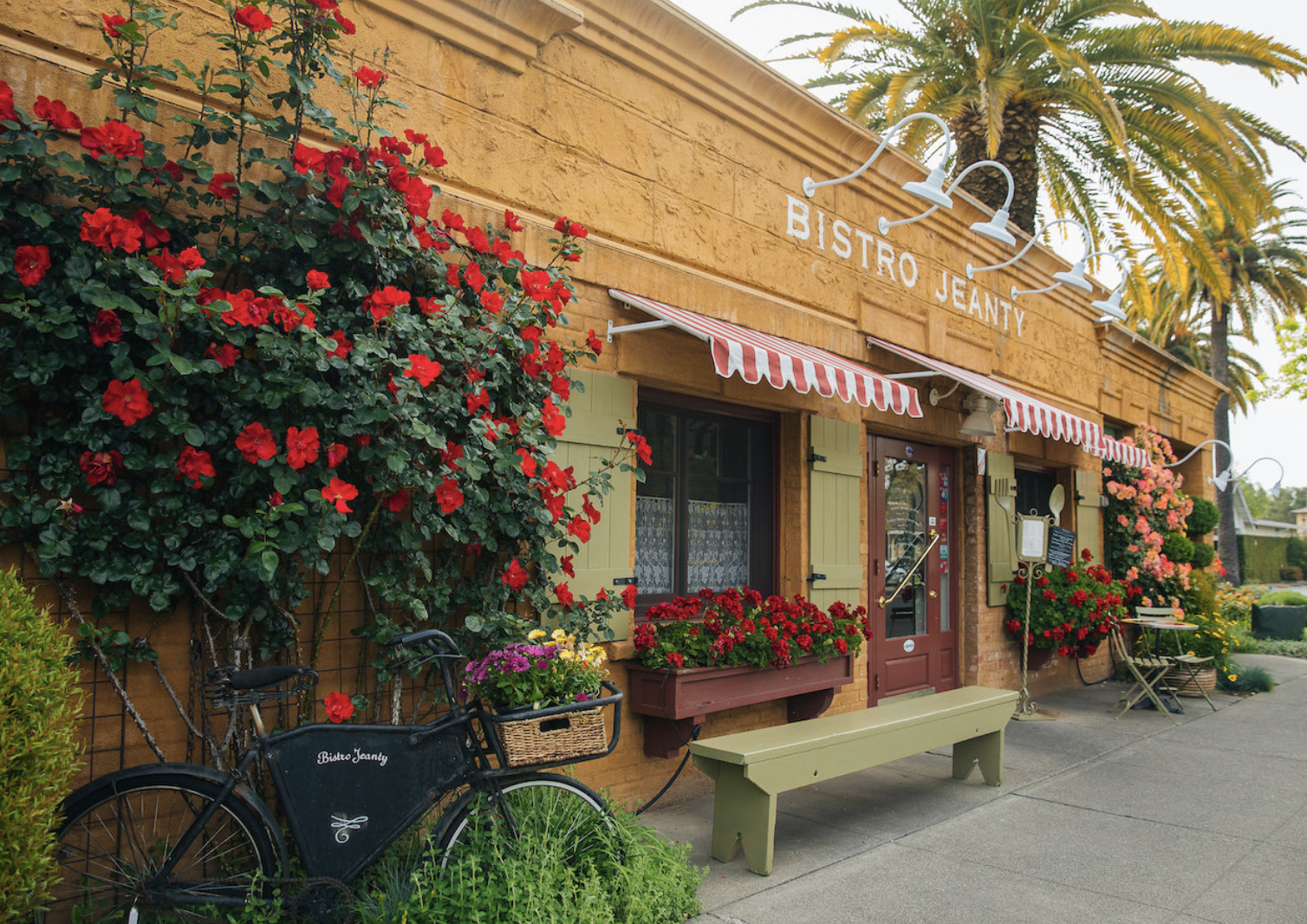 Exterior of Bistro Jeanty in Yountville, Napa Valley, featuring colorful rose gardens, striped awnings, palm trees, and a charming French bistro entrance.