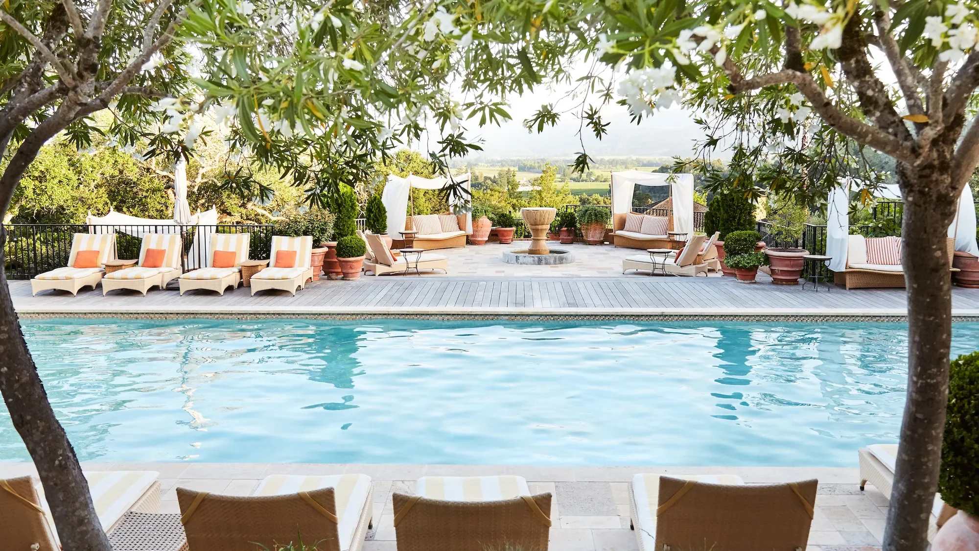 Luxury resort pool in Napa Valley surrounded by cabanas, lounge chairs, mature trees, and landscaped gardens with vineyard views in the distance.
