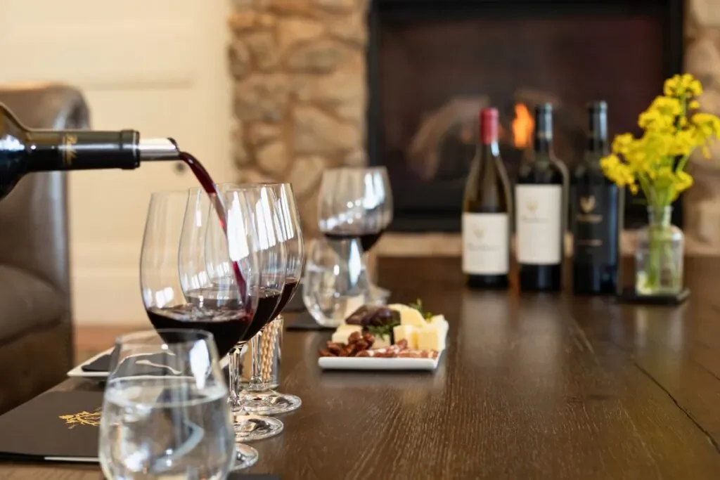 Red wine being poured into tasting glasses during a private wine tasting in Napa Valley, with bottles, a charcuterie plate, and a cozy fireplace in the background.