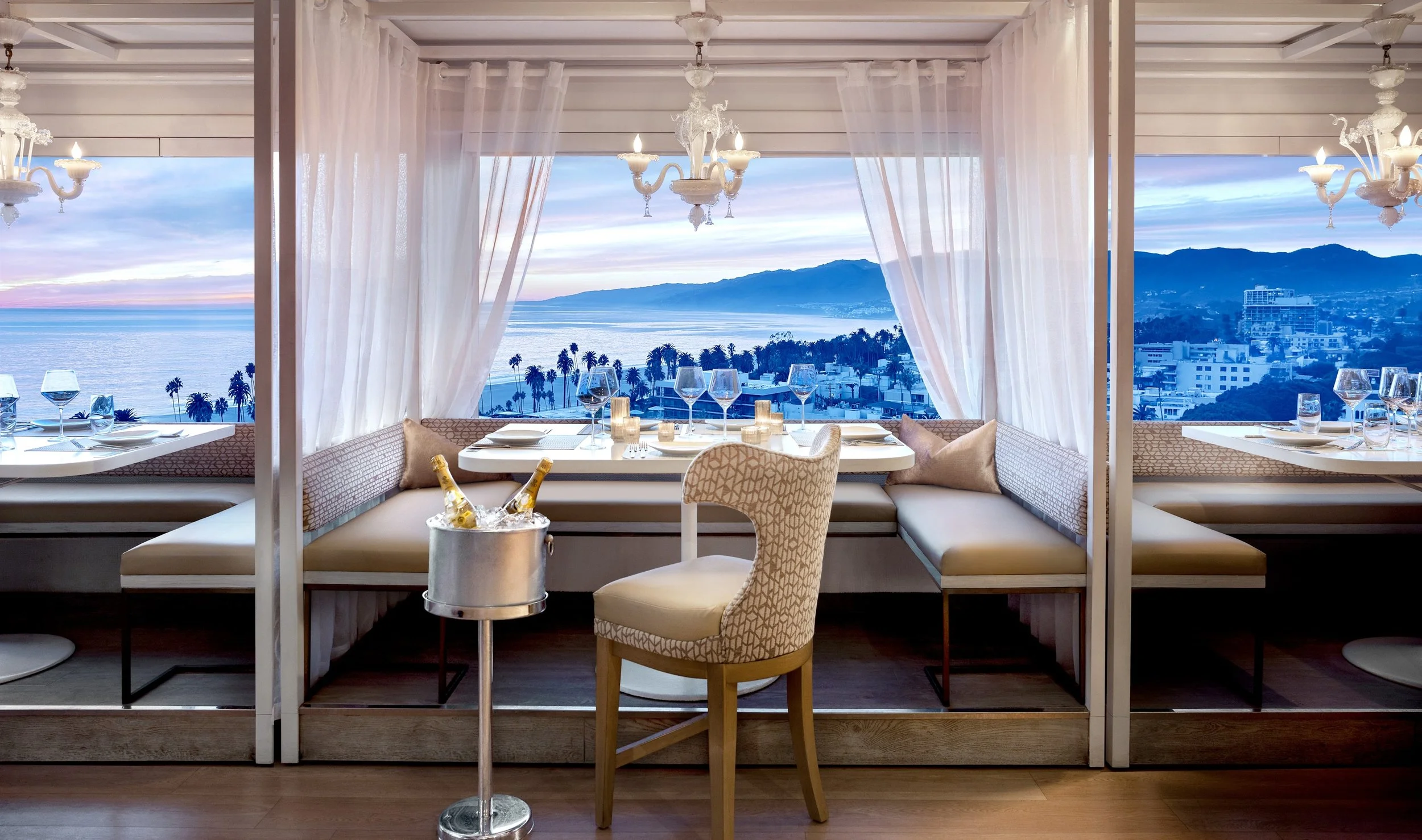 An elegant oceanfront dining room overlooks the Santa Monica coastline at sunset, featuring intimate booth seating, soft drapery, candlelit tables, and sweeping views of the Pacific Ocean and California mountains.