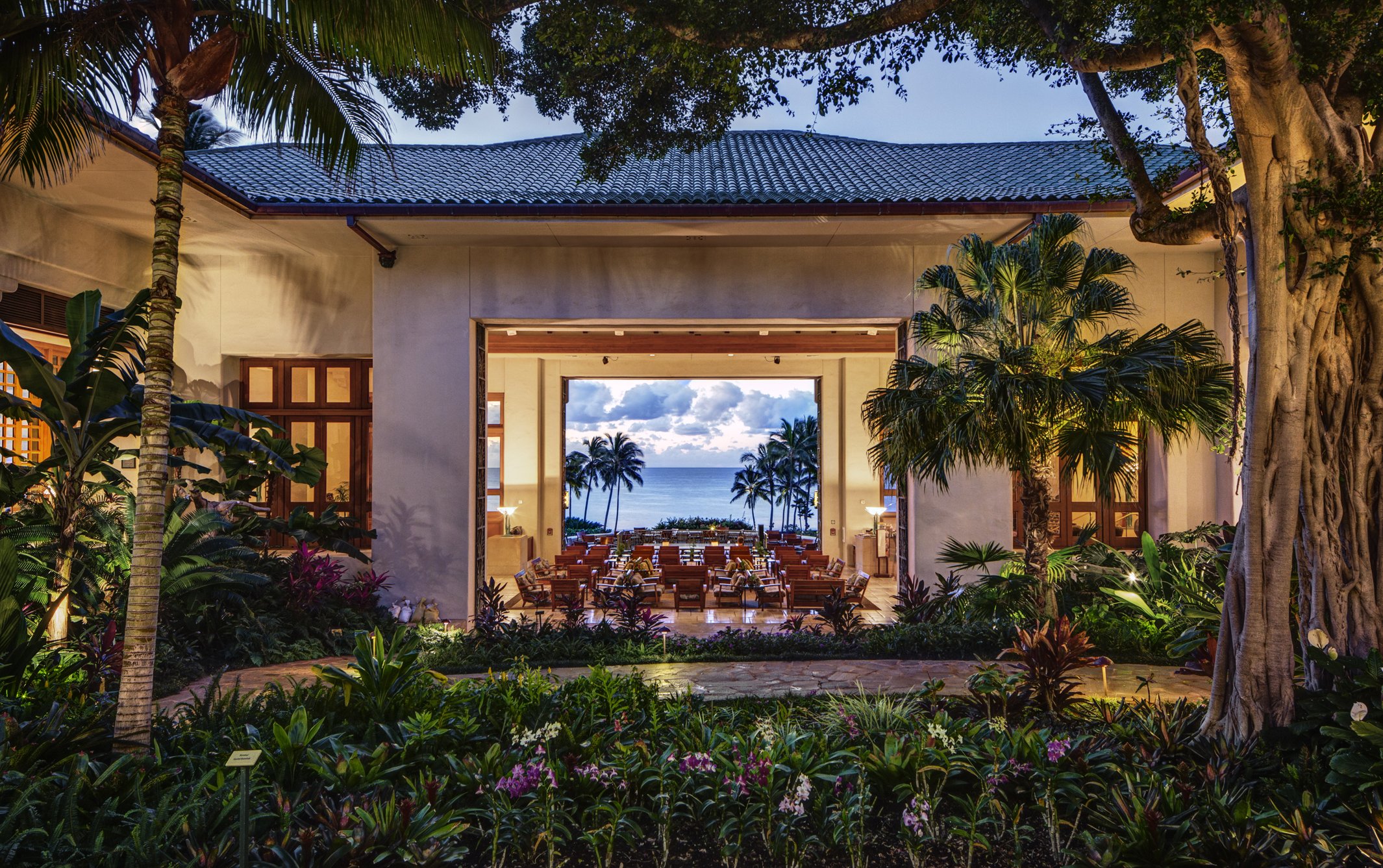 Open-air lobby at Grand Hyatt Kauai Resort & Spa in Poipu, Hawaii overlooking palm trees and the Pacific Ocean at sunset.