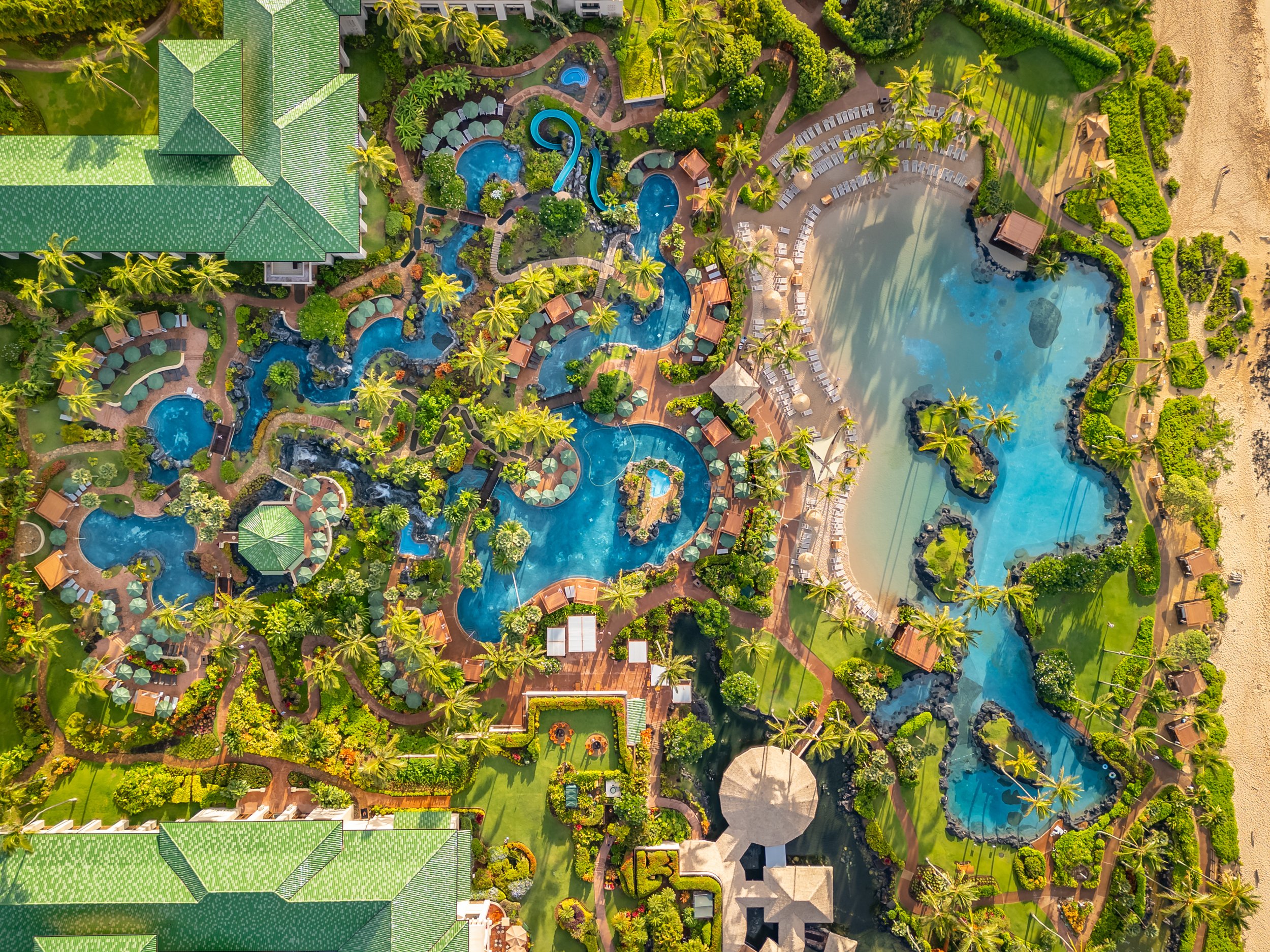 Top-down aerial view of Grand Hyatt Kauai Resort & Spa pools featuring winding lagoon pools, waterfalls, tropical gardens, and Poipu Beach coastline.