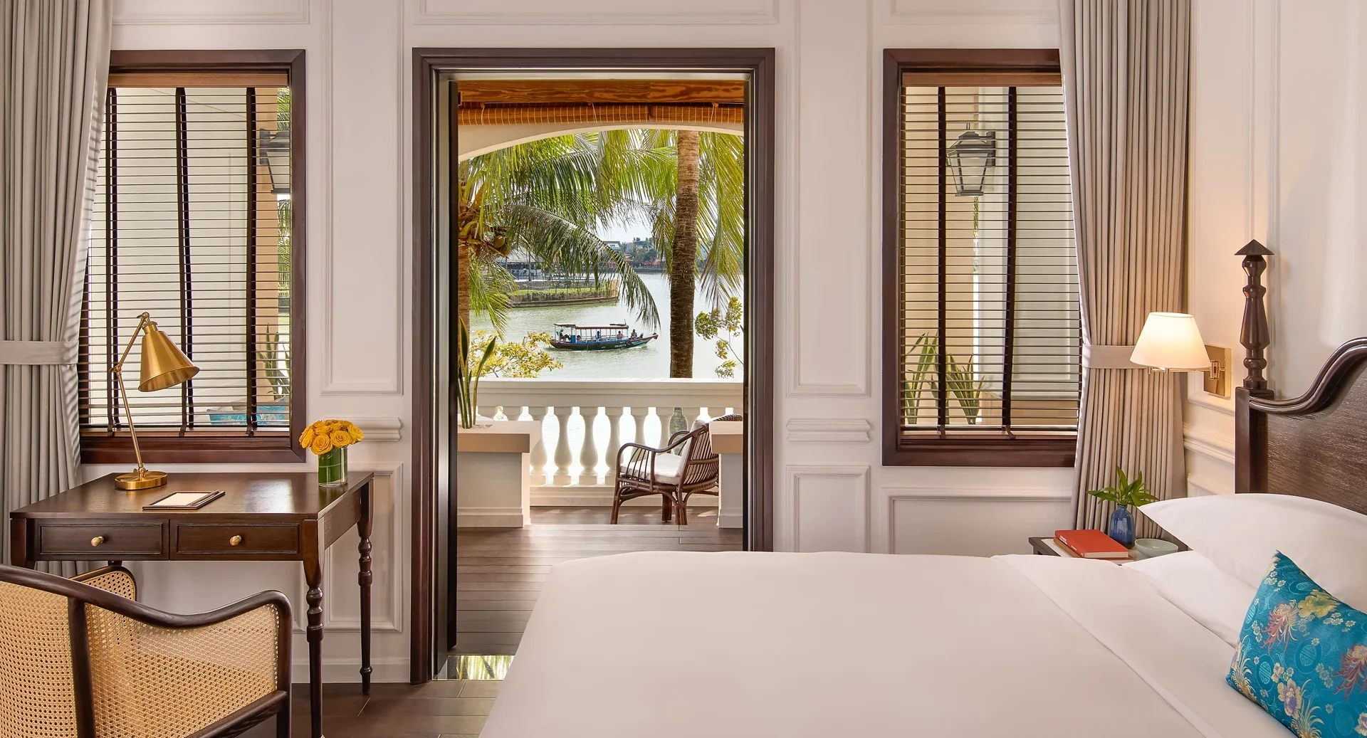 Elegant room at Anantara Hoi An Resort featuring a plush bed, French colonial design, and private balcony overlooking the river with traditional Vietnamese boat.