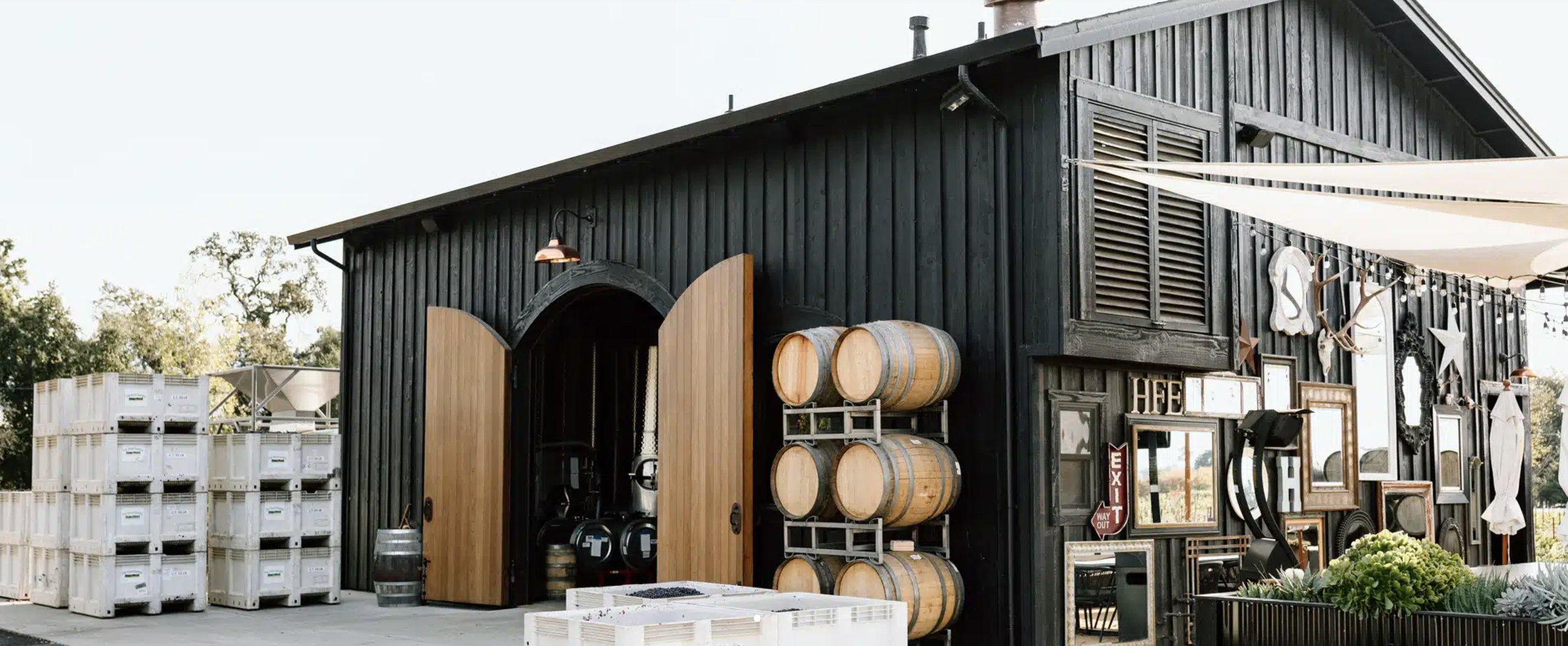 Modern black winery building in Napa Valley with open wooden doors, stacked wine barrels, and outdoor décor, showcasing a contemporary tasting room and wine production facility.