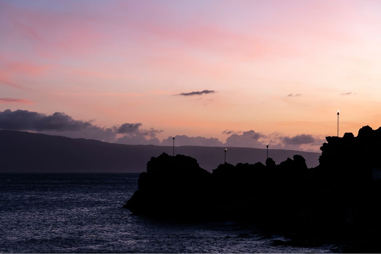 Kaanapali Maui black rock sunset with ocean view and rocky coastline silhouetted against a soft pink and purple sky.