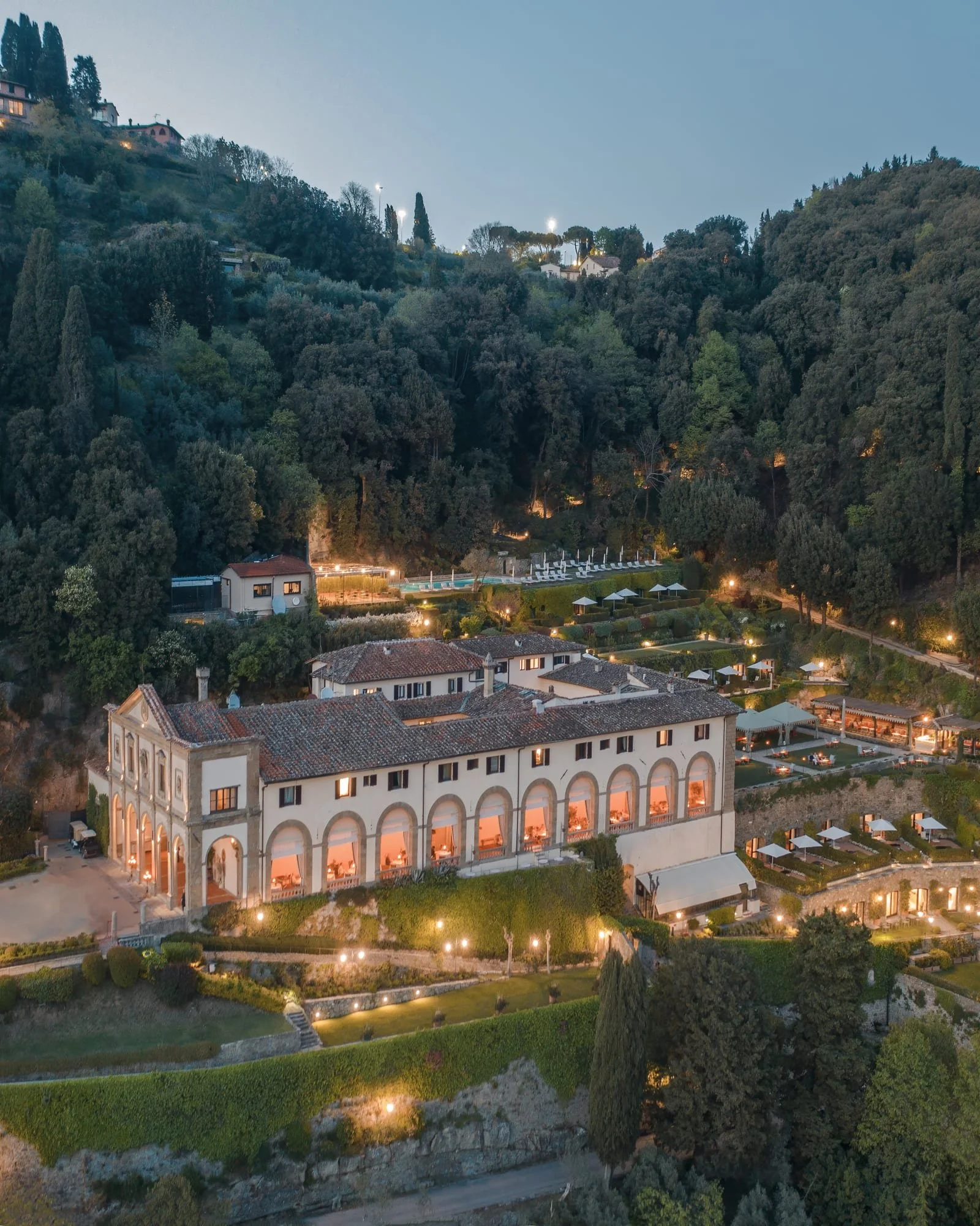 Aerial view of a luxury historic villa hotel in the hills of Florence, Italy, surrounded by lush gardens, terraces, and trees at dusk.