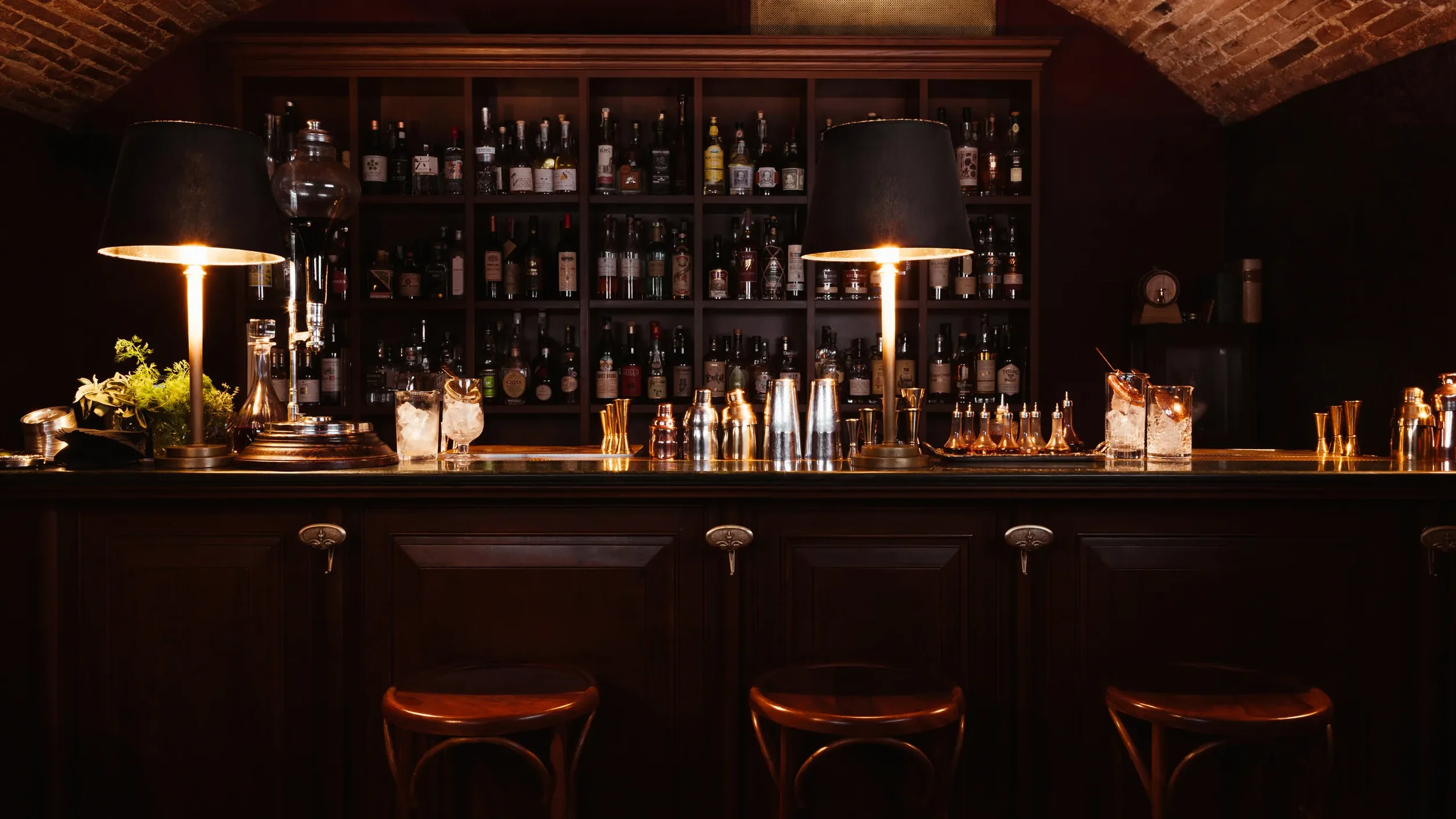 Elegant, dimly lit cocktail bar with dark wood interiors, glowing table lamps, and shelves filled with spirits.