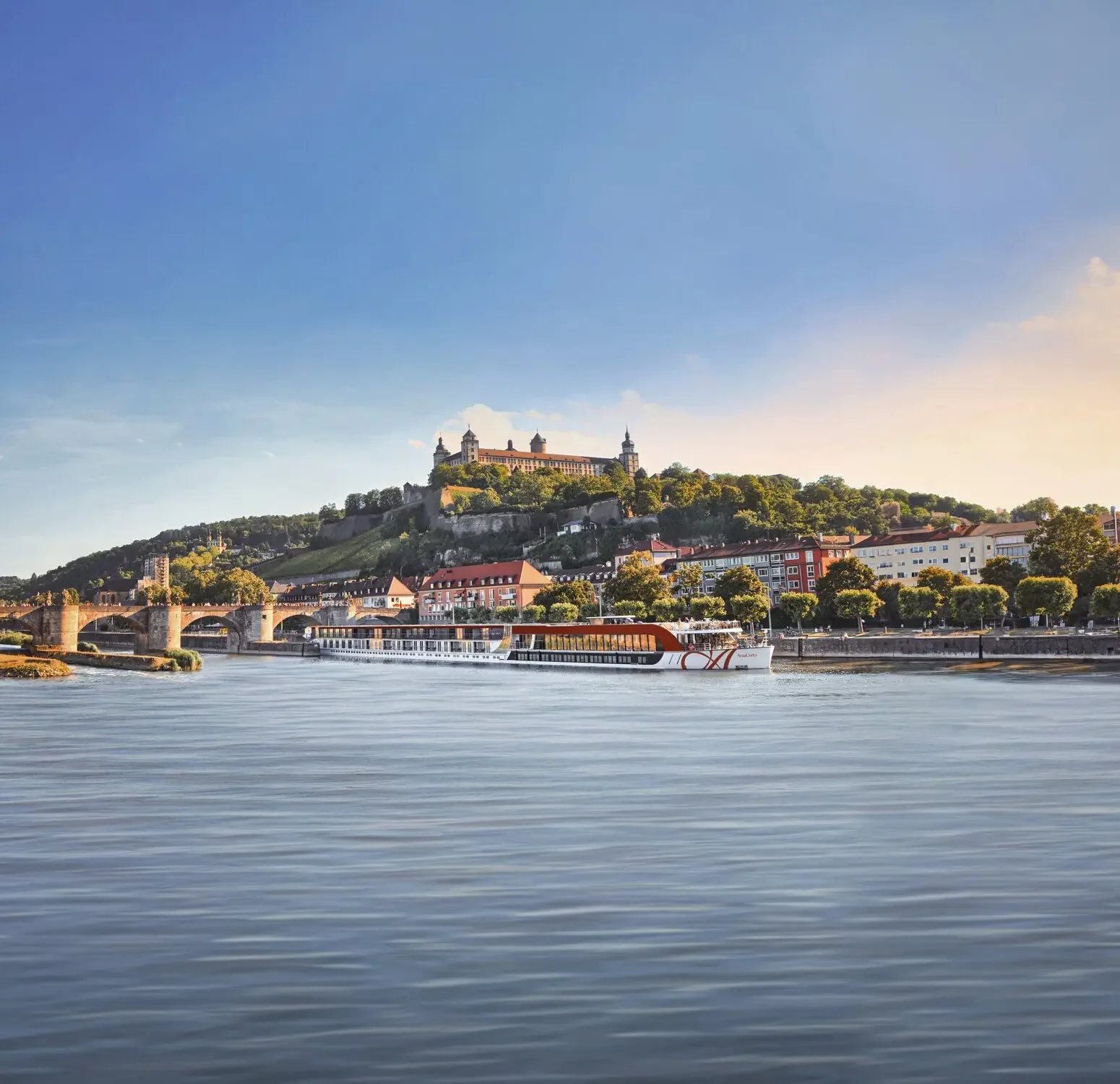 Scenic view of Marienberg Fortress overlooking Würzburg, Germany, with a luxury river cruise ship sailing along the Main River beneath a clear blue sky.