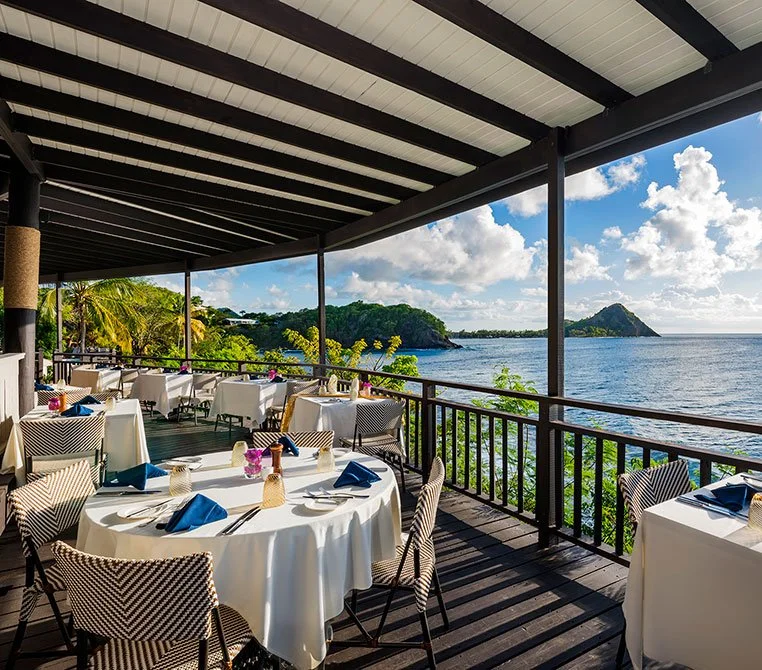 Luxury open-air cliffside restaurant in St. Lucia featuring elegant waterfront dining, white tablecloths, and panoramic views of the Caribbean Sea and lush tropical coastline.