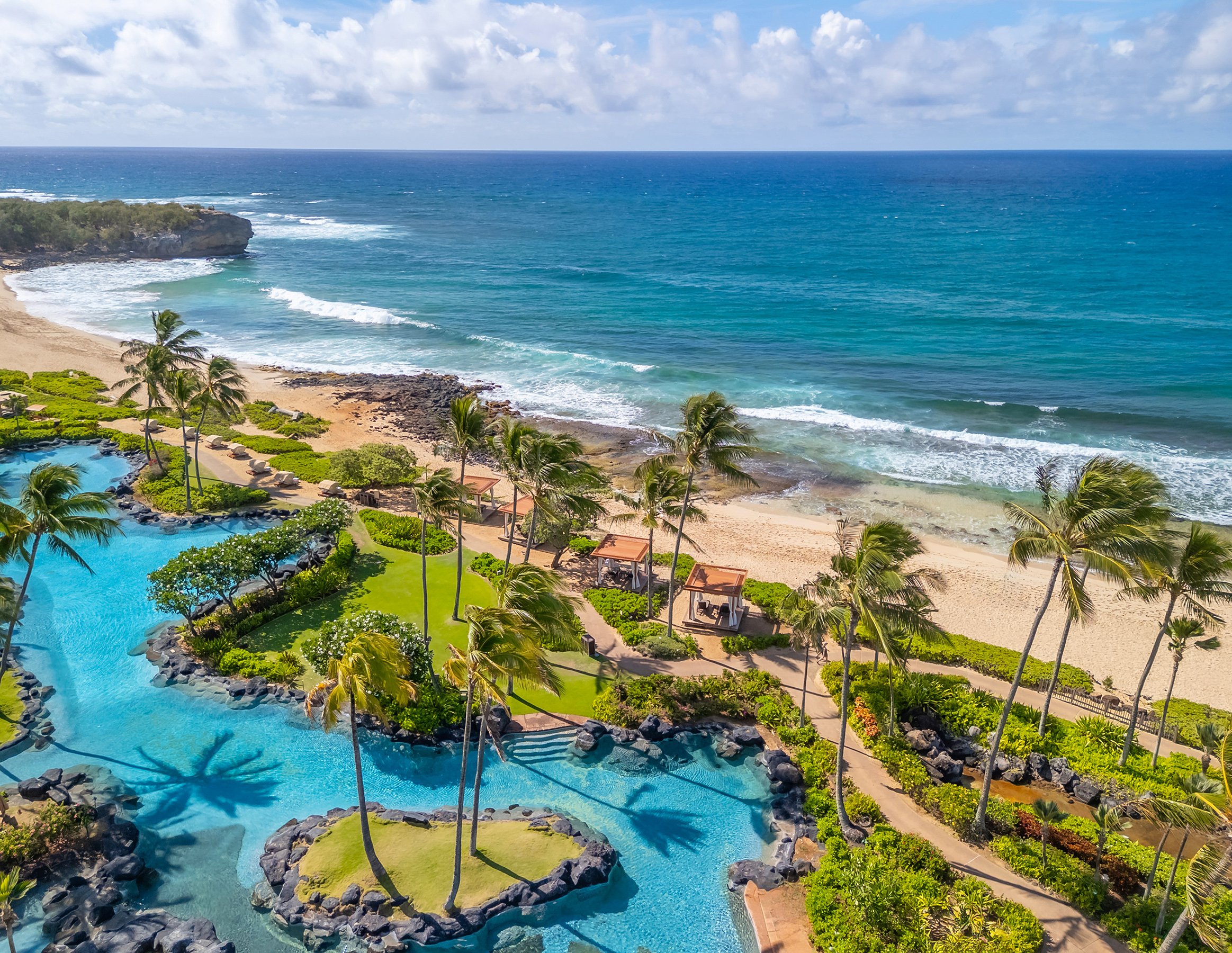 Oceanfront lagoon pools at Grand Hyatt Kauai Resort & Spa overlooking Poipu Beach with palm trees and the Pacific Ocean.