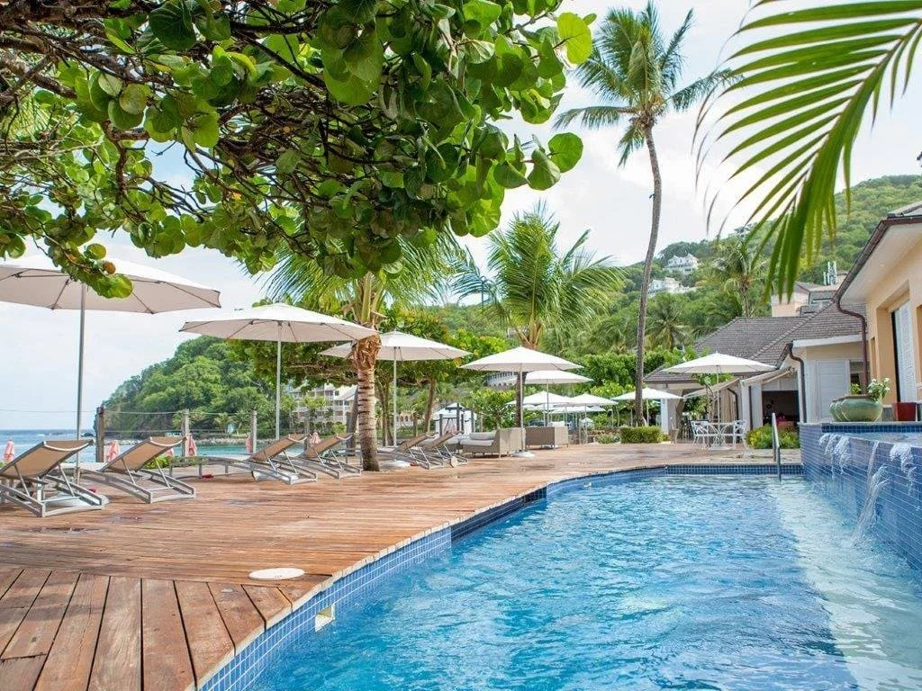 Luxury beachfront resort in St. Lucia featuring a swimming pool, wooden sundeck, lounge chairs, white umbrellas, and lush tropical gardens overlooking the Caribbean Sea.
