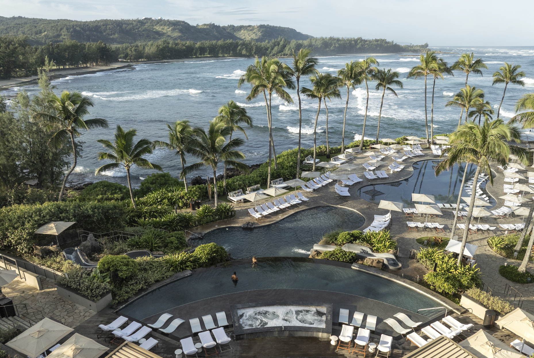 Aerial view of The Ritz-Carlton Turtle Bay pools with oceanfront infinity pool, palm trees, and panoramic North Shore views.