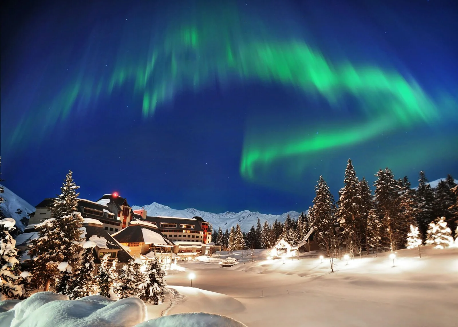Northern Lights glowing over a snowy lodge in Alaska with pine trees and mountains under a night sky