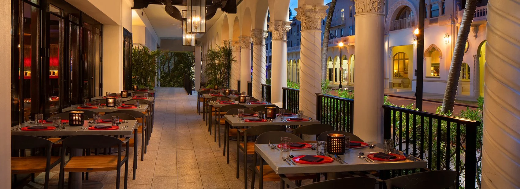 Elegant outdoor restaurant terrace at a luxury hotel featuring candlelit dining tables, ornate white columns, tropical greenery, and Mediterranean-inspired architecture at dusk.