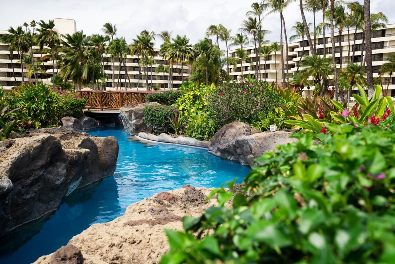 Beautiful tropical resort lagoon pool in Maui with winding waterways, lush gardens, palm trees, and a wooden bridge connecting landscaped pathways.